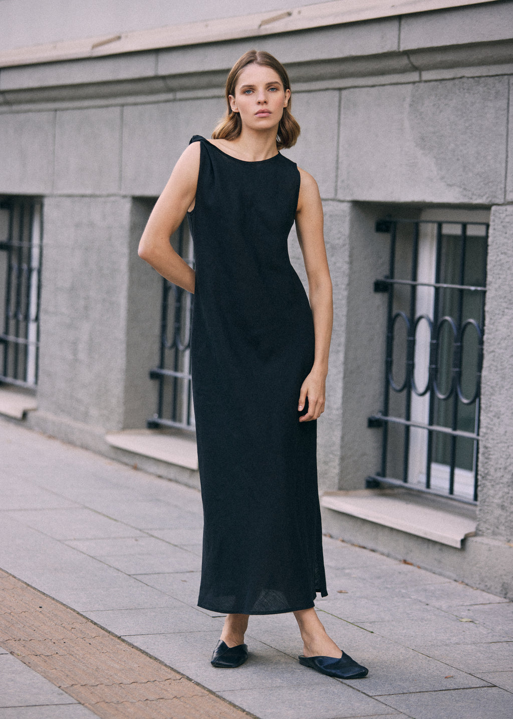 Woman wearing a long black sleeveless linen maxi dress with a clean, minimalist silhouette, styled with black flat shoes on a city street.