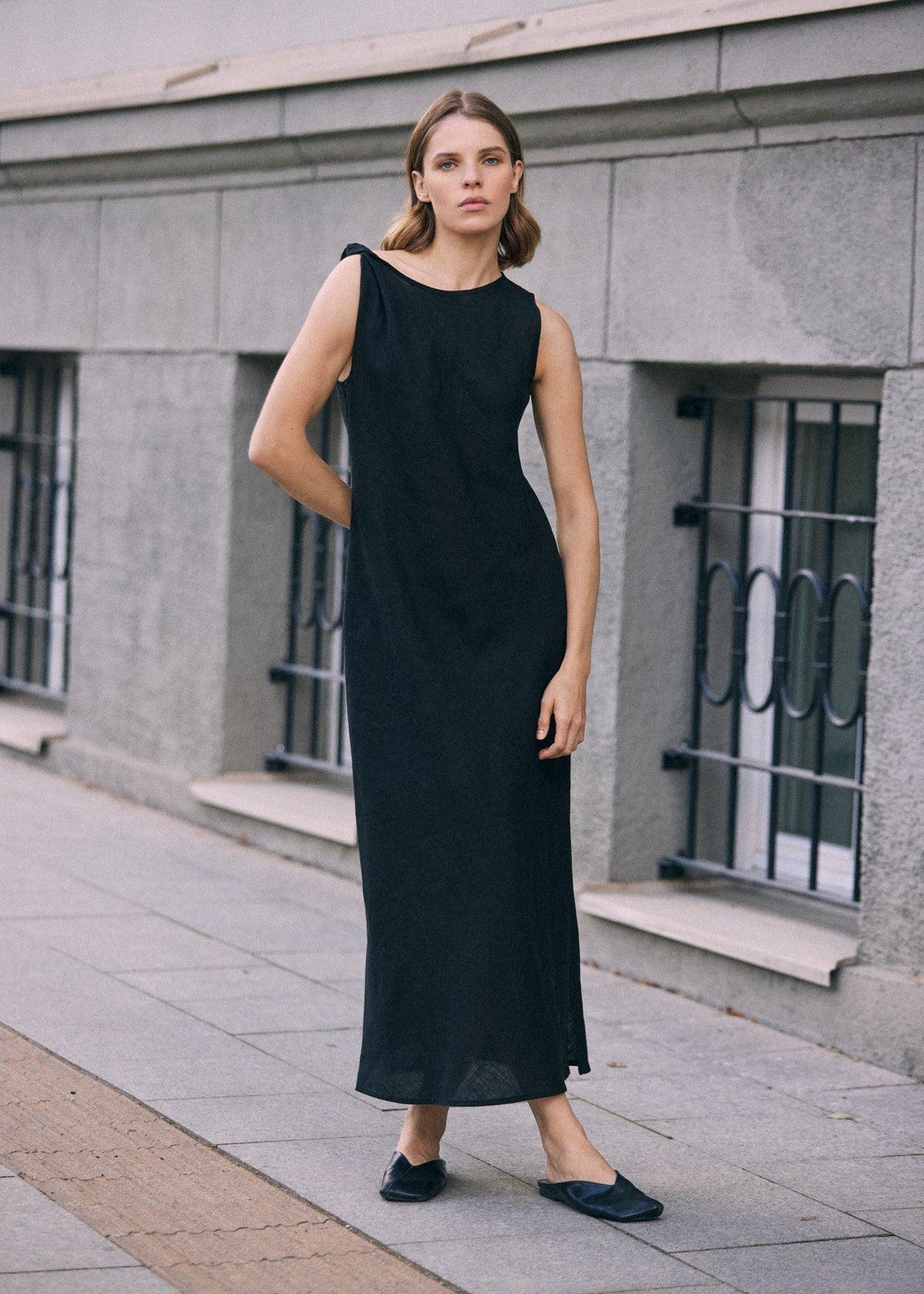 Woman wearing a long black sleeveless linen maxi dress with a clean, minimalist silhouette, styled with black flat shoes on a city street.