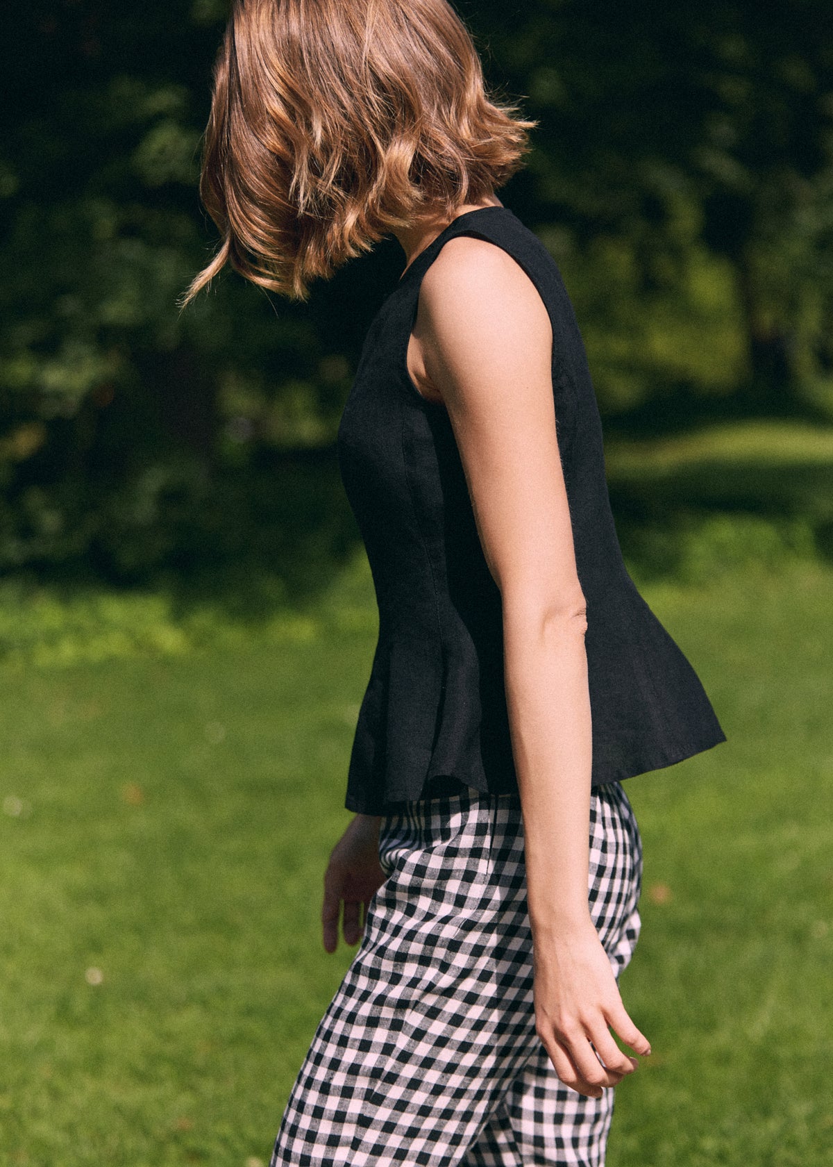 A woman wearing a sleeveless black peplum top and black-and-white gingham trousers, photographed outdoors on a grassy lawn with trees in the background.