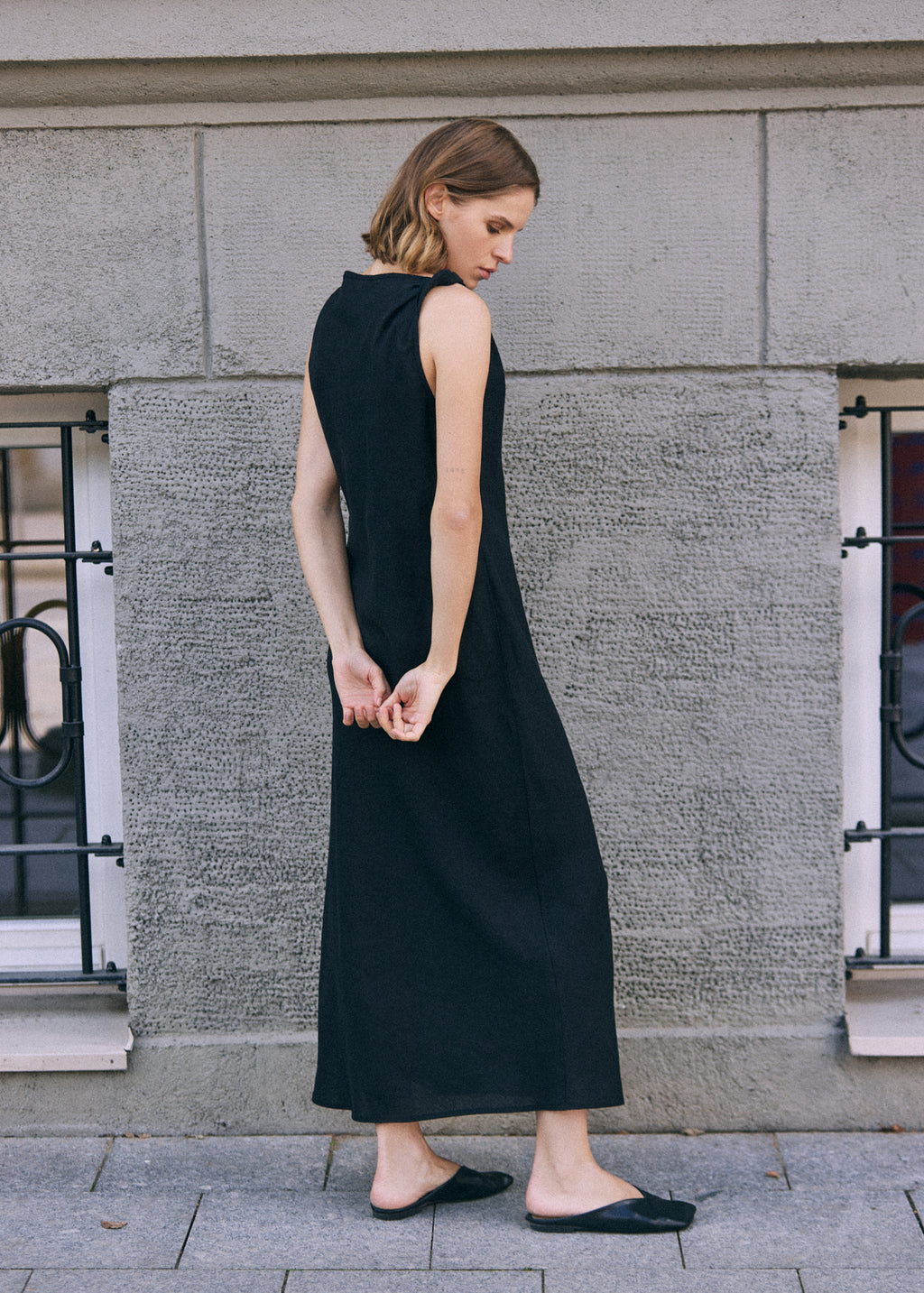Woman shown from the back wearing a long black sleeveless linen dress with a clean, flowing silhouette and twisted shoulder detail, styled with black flat shoes in an urban setting.