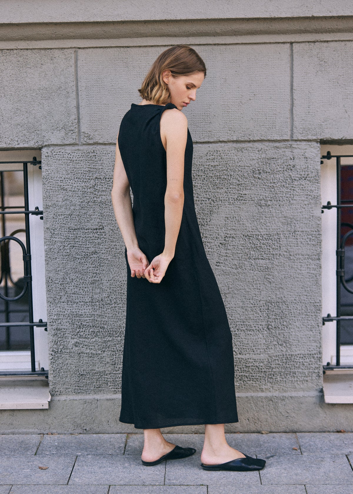 Woman shown from the back wearing a long black sleeveless linen dress with a clean, flowing silhouette and twisted shoulder detail, styled with black flat shoes in an urban setting.