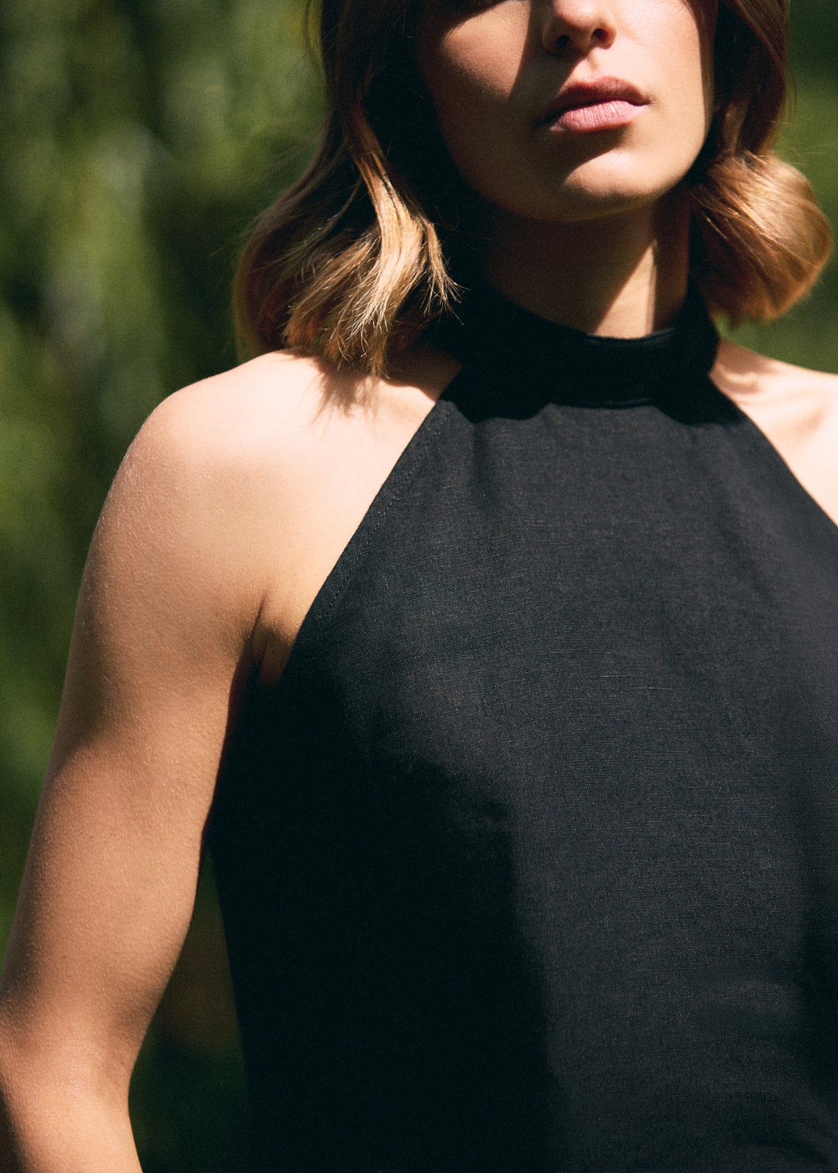 A close-up of a woman wearing a black halter-neck top, photographed outdoors with natural sunlight and a blurred green background.