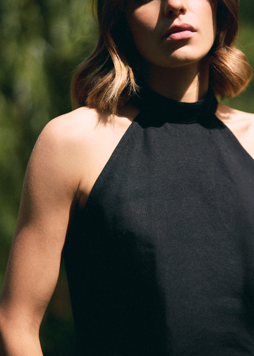 A close-up of a woman wearing a black halter-neck top, photographed outdoors with natural sunlight and a blurred green background.