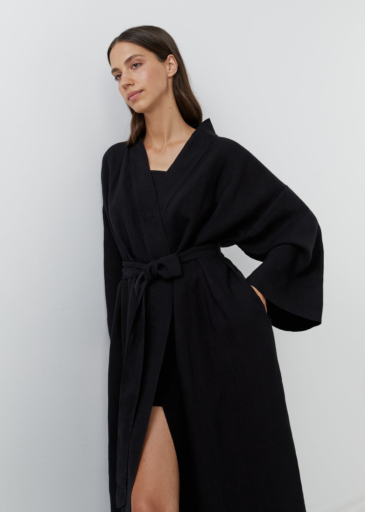 A woman wearing a long black linen wrap robe with wide sleeves and a self-tie belt, standing against a plain light gray wall.