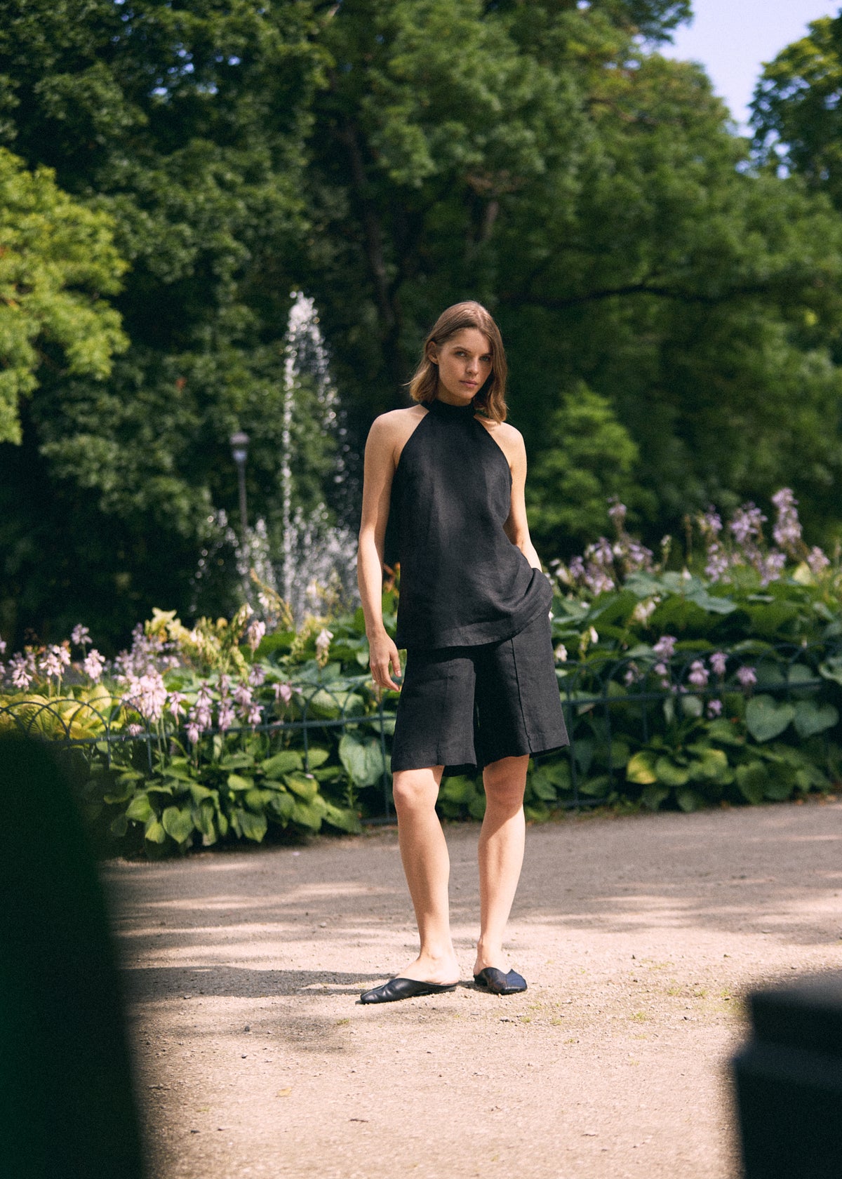 A woman stands outdoors in a park-like setting with trees, flowers, and a path, wearing a black sleeveless high-neck top and matching black shorts  with black flat shoes.