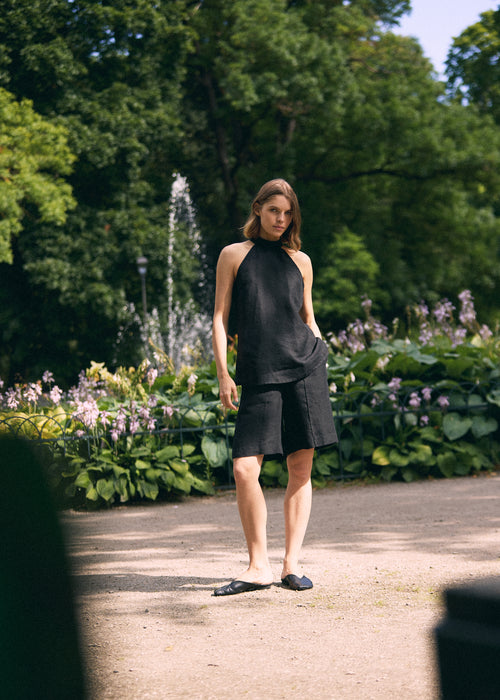 A woman stands outdoors in a park-like setting with trees, flowers, and a path, wearing a black sleeveless high-neck top and matching black shorts  with black flat shoes.