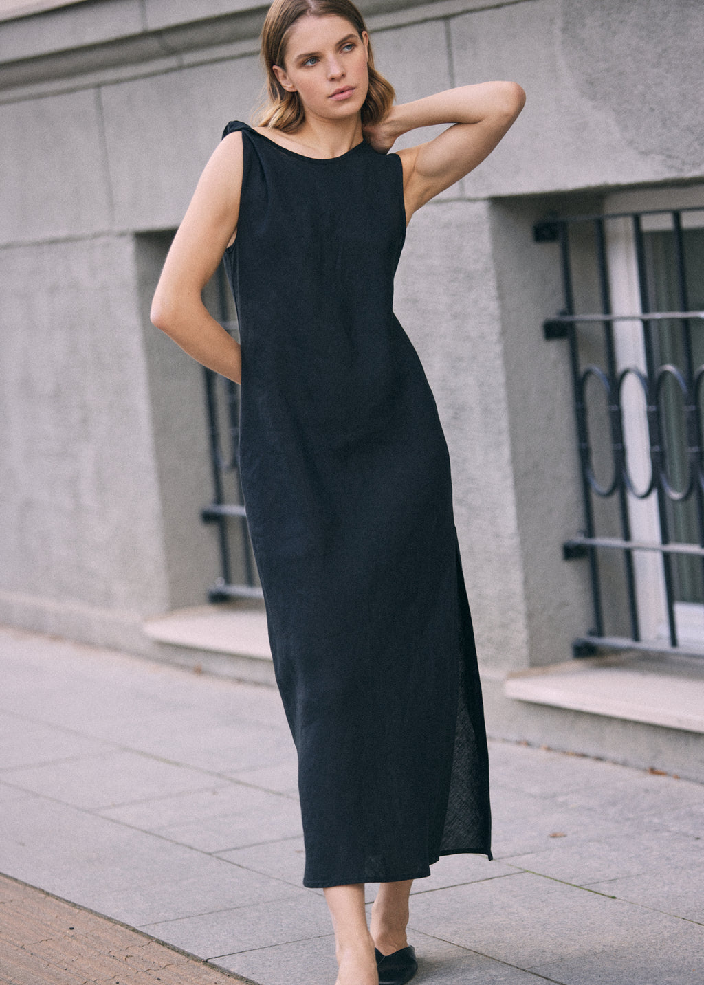 Woman wearing a black sleeveless linen maxi dress with a subtle twisted shoulder detail and a softly fitted silhouette, standing against a grey urban building.