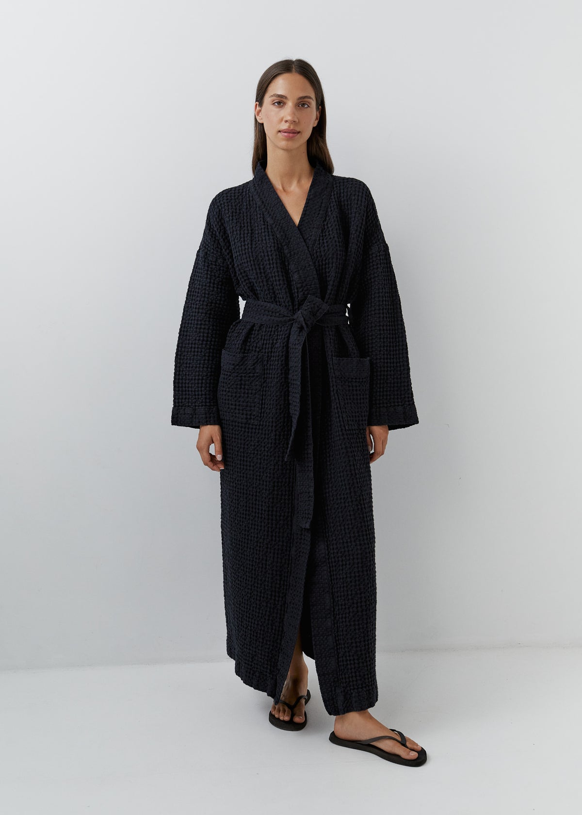 A woman modeling a long black waffle-textured bathrobe with a tie waist and front pockets, standing against a plain light gray studio background.