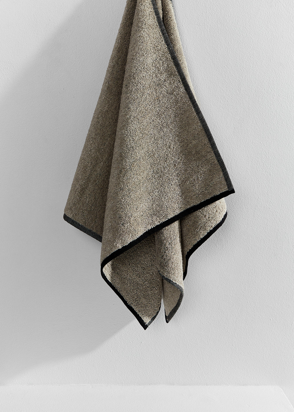 A textured sand-coloured linen towel with contrasting black trim, hanging against a plain white wall.