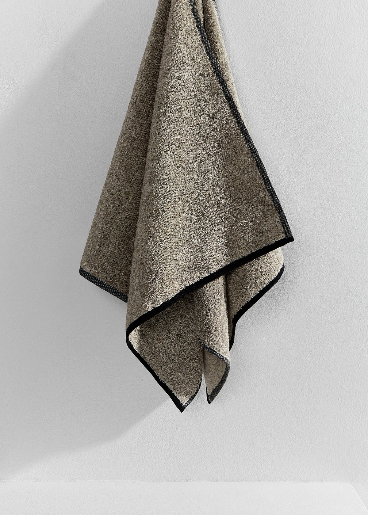 A textured sand-coloured linen towel with contrasting black trim, hanging against a plain white wall.
