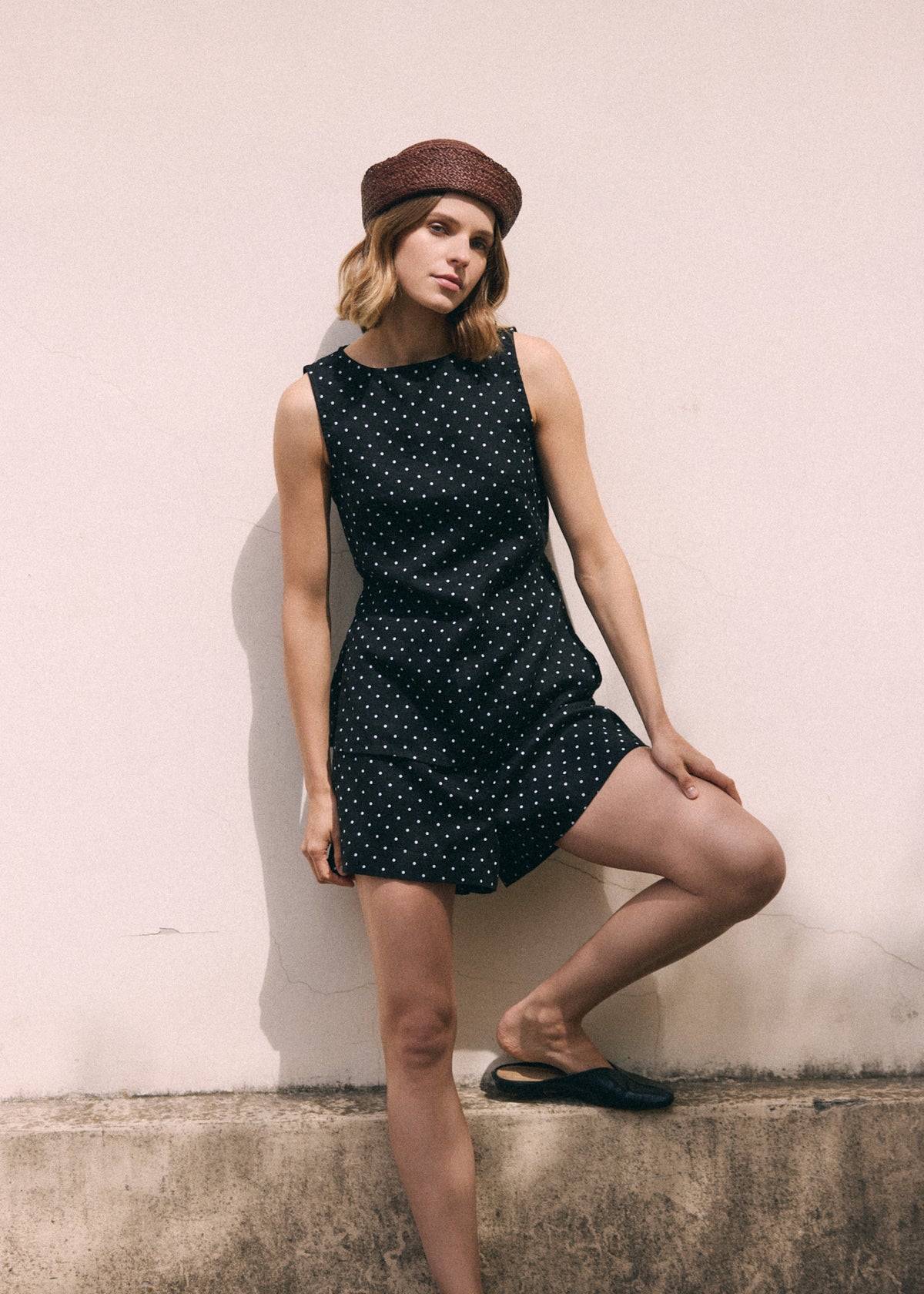 A woman poses wearing a black sleeveless top with white polka dots and matching shorts, a brown beret, and black flat shoes against a textured light pink wall.