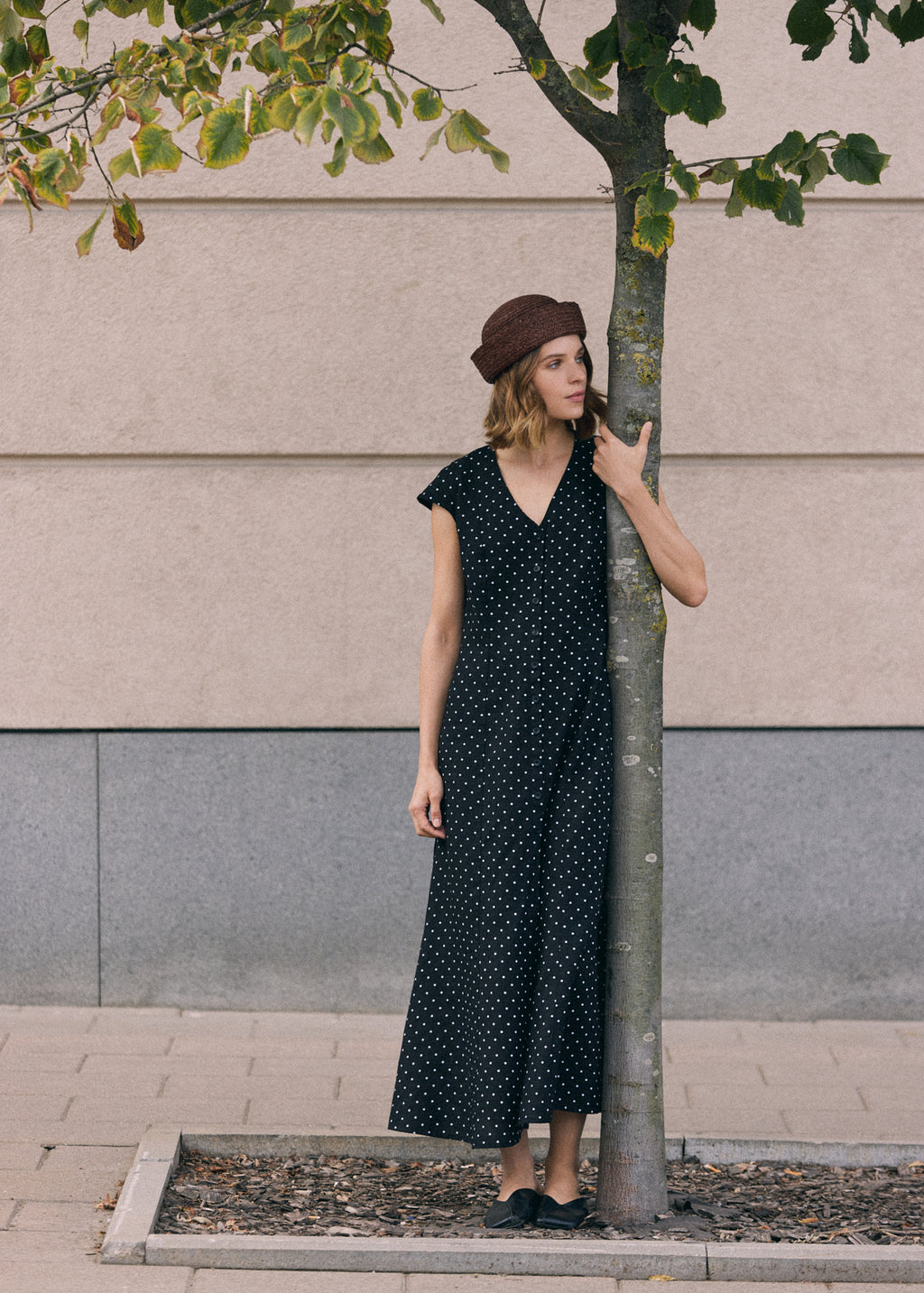 Woman wearing a black linen maxi dress with white polka dots, V-neckline, short cap sleeves and front button closure, standing outdoors by a tree with a brown hat and black shoes.