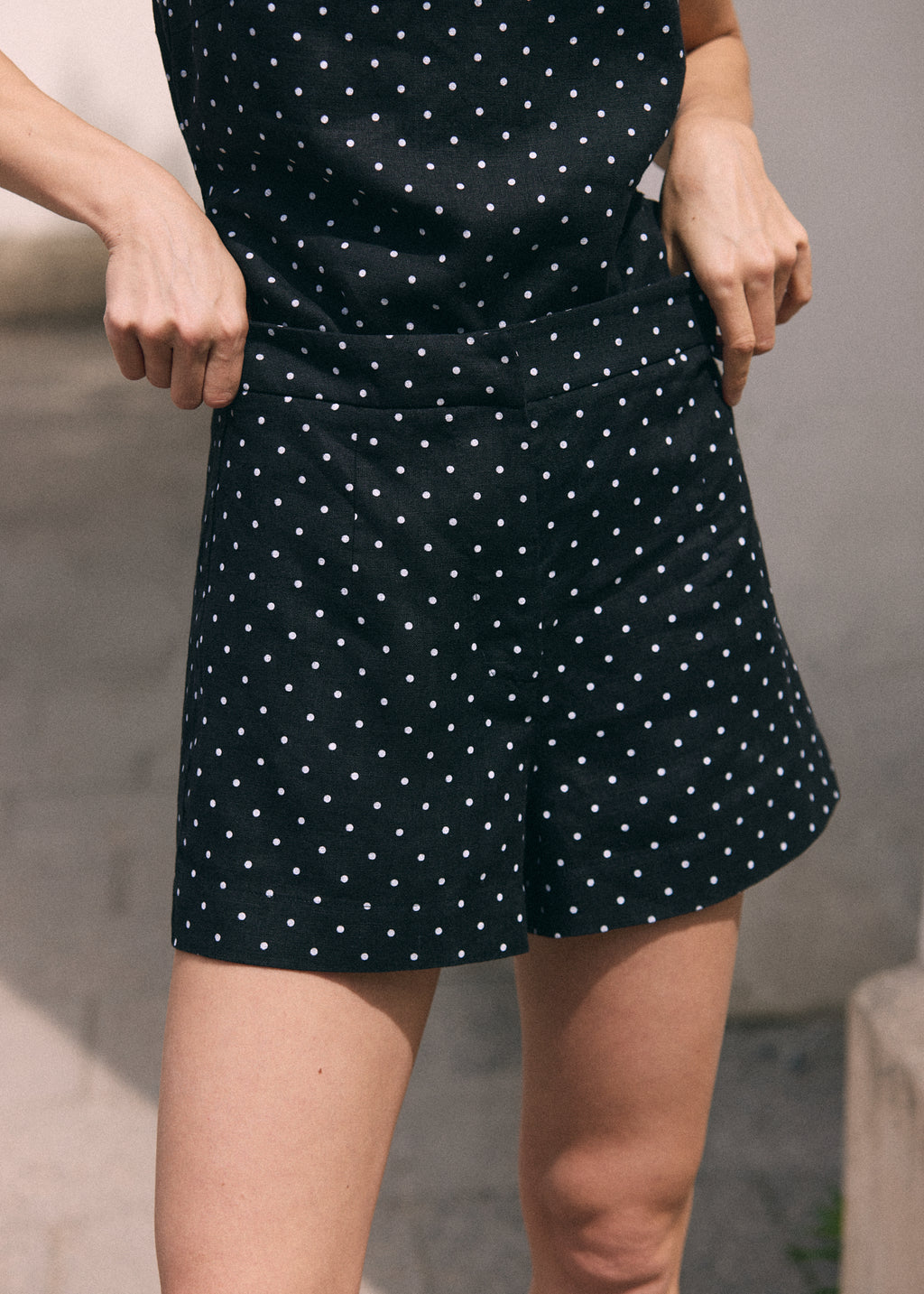 A close-up of a person wearing black shorts with a white polka dot pattern, holding the sides of the shorts to show their shape and fit.