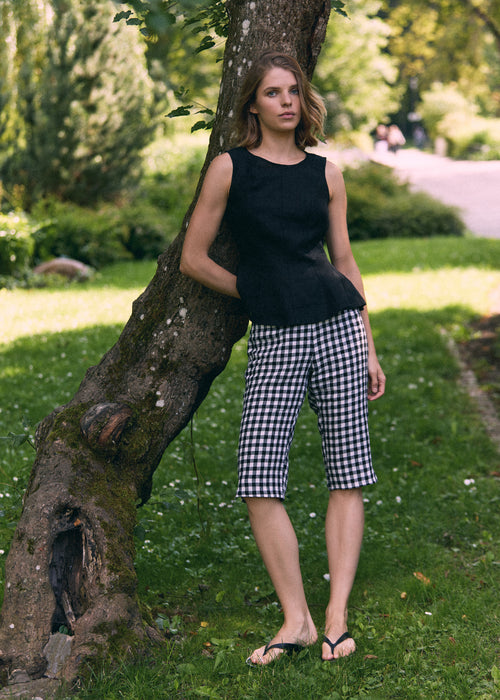 A woman stands outdoors leaning against a tree on a grassy path, wearing a sleeveless black top and black-and-white gingham capri pants with black flip-flops.