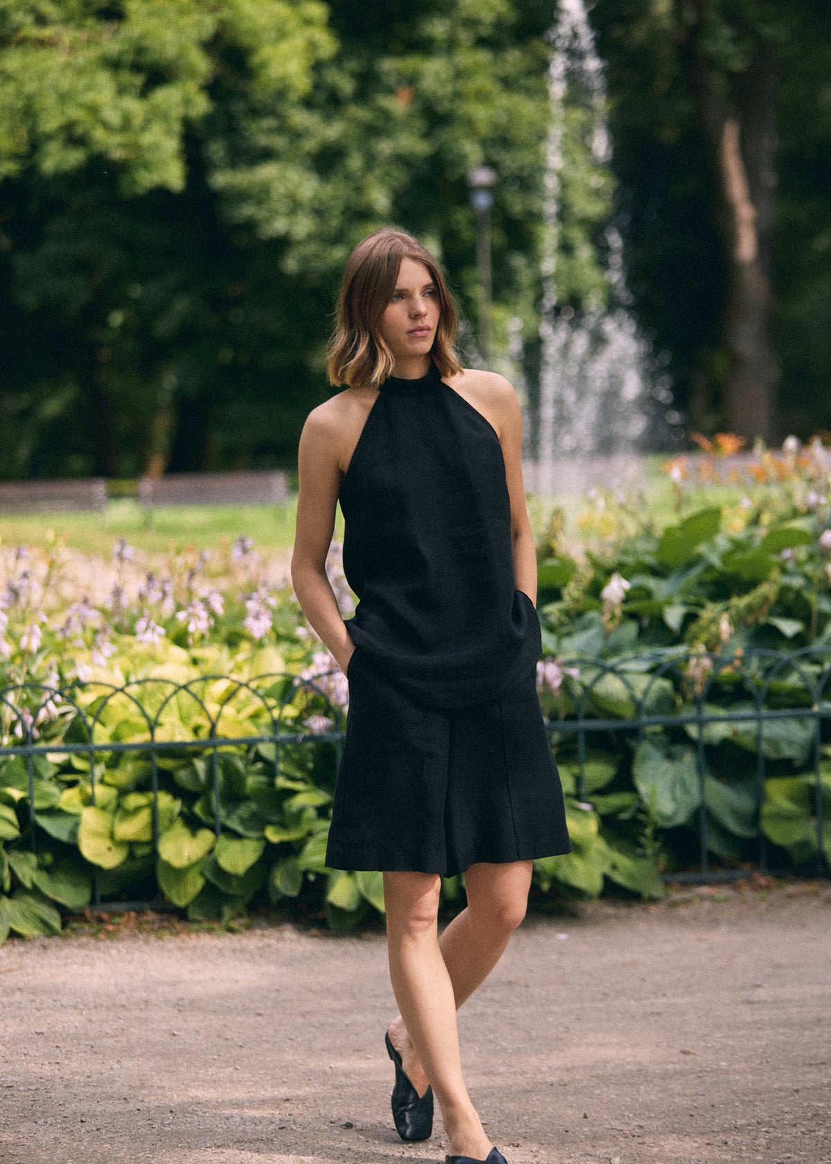 A woman wearing black knee-length shorts and a black sleeveless top walks along a park path with a fountain and greenery in the background.