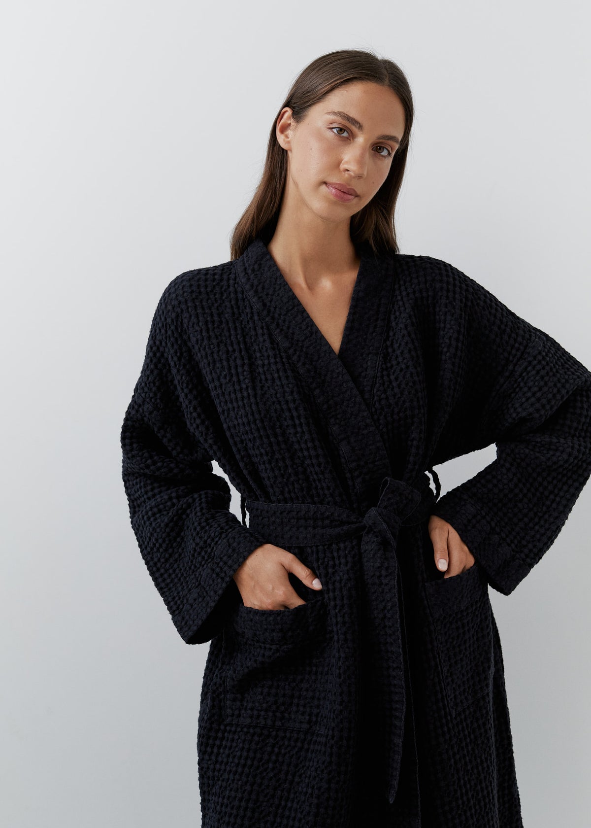 A woman wearing a black waffle linen robe with a tie belt, standing against a light gray studio background.
