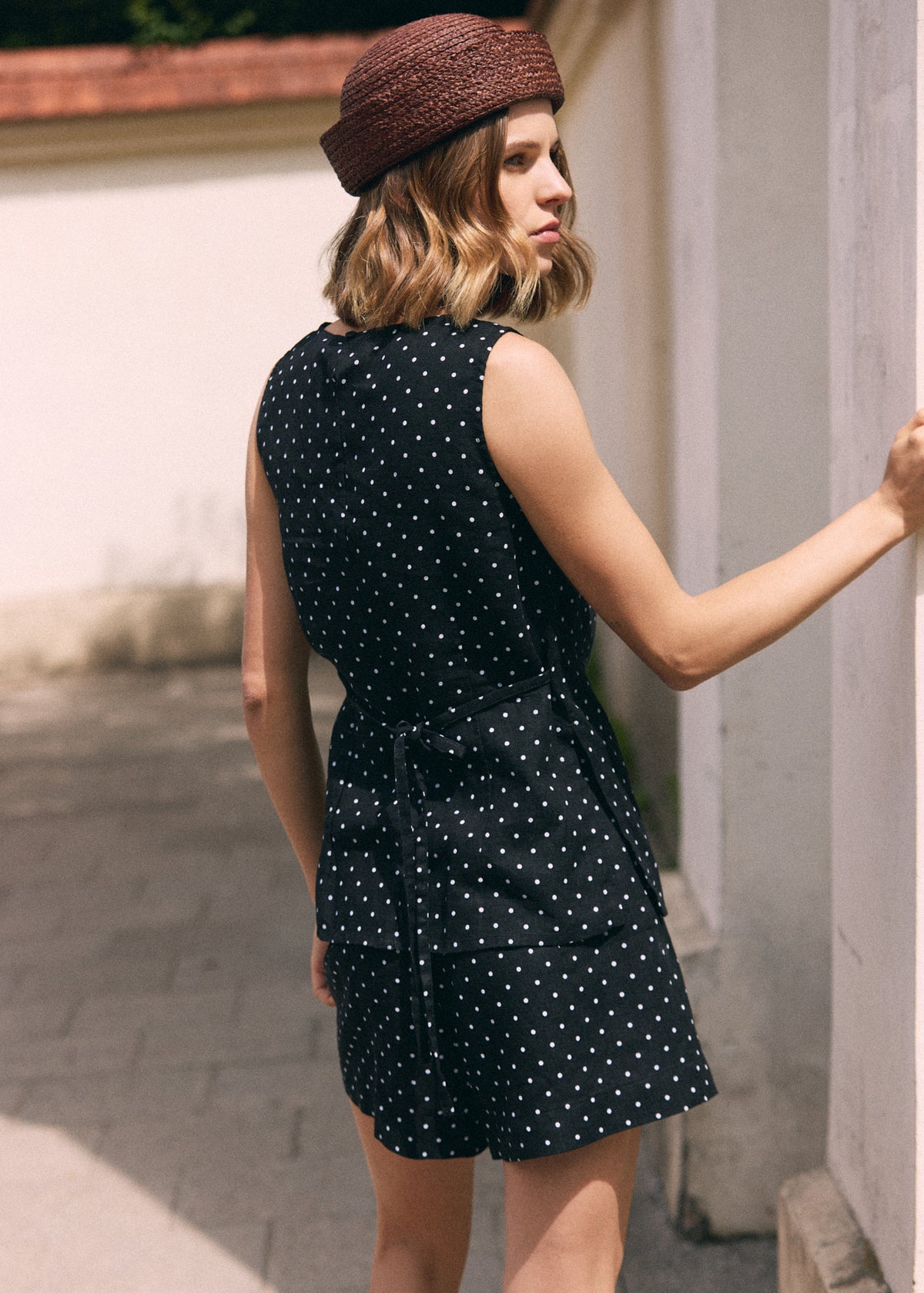 A woman wearing a black sleeveless top with white polka dots and matching shorts, paired with a brown textured beret, photographed outdoors against a light-colored wall.
