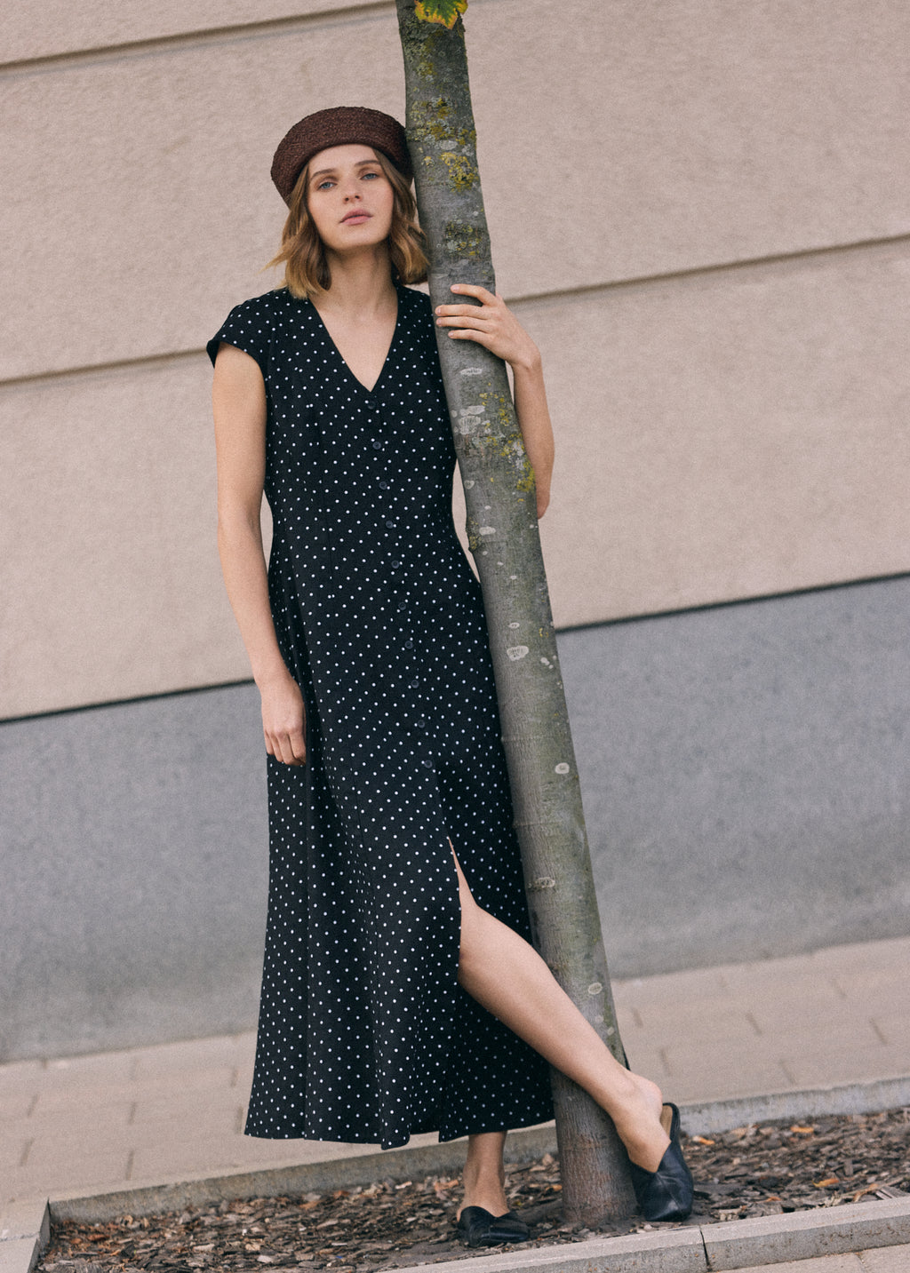Woman wearing a black linen maxi dress with white polka dot pattern, V-neckline, short sleeves, front button closure and side slit, standing by a tree outdoors and wearing a brown beret and black shoes.