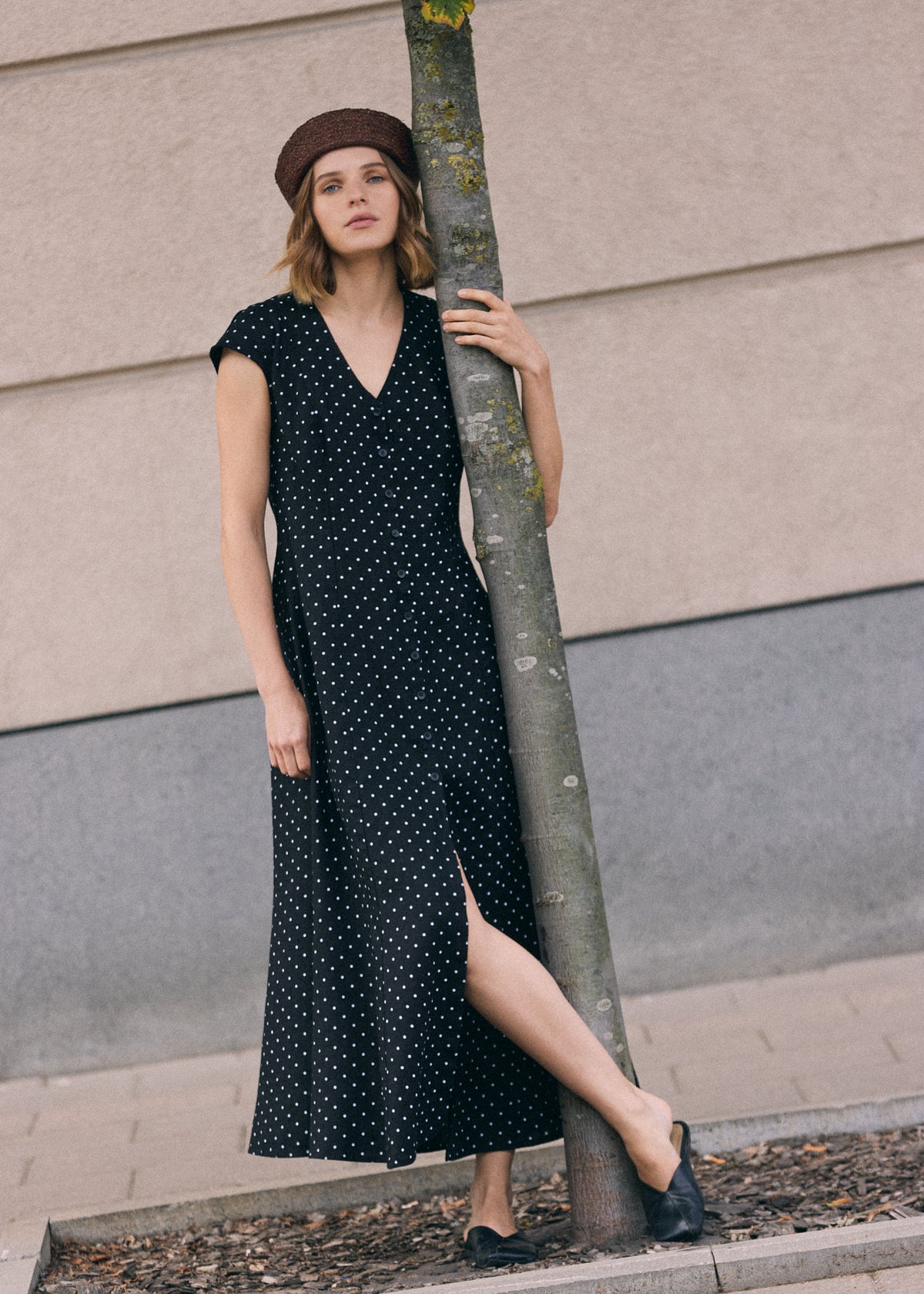 Woman wearing a black linen maxi dress with white polka dot pattern, V-neckline, short sleeves, front button closure and side slit, standing by a tree outdoors and wearing a brown beret and black shoes.