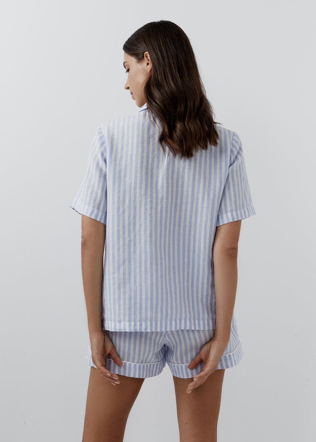 A woman shown from the back wearing a light blue and white vertically striped short-sleeve linen pajama top and matching linen shorts against a plain light gray studio background.