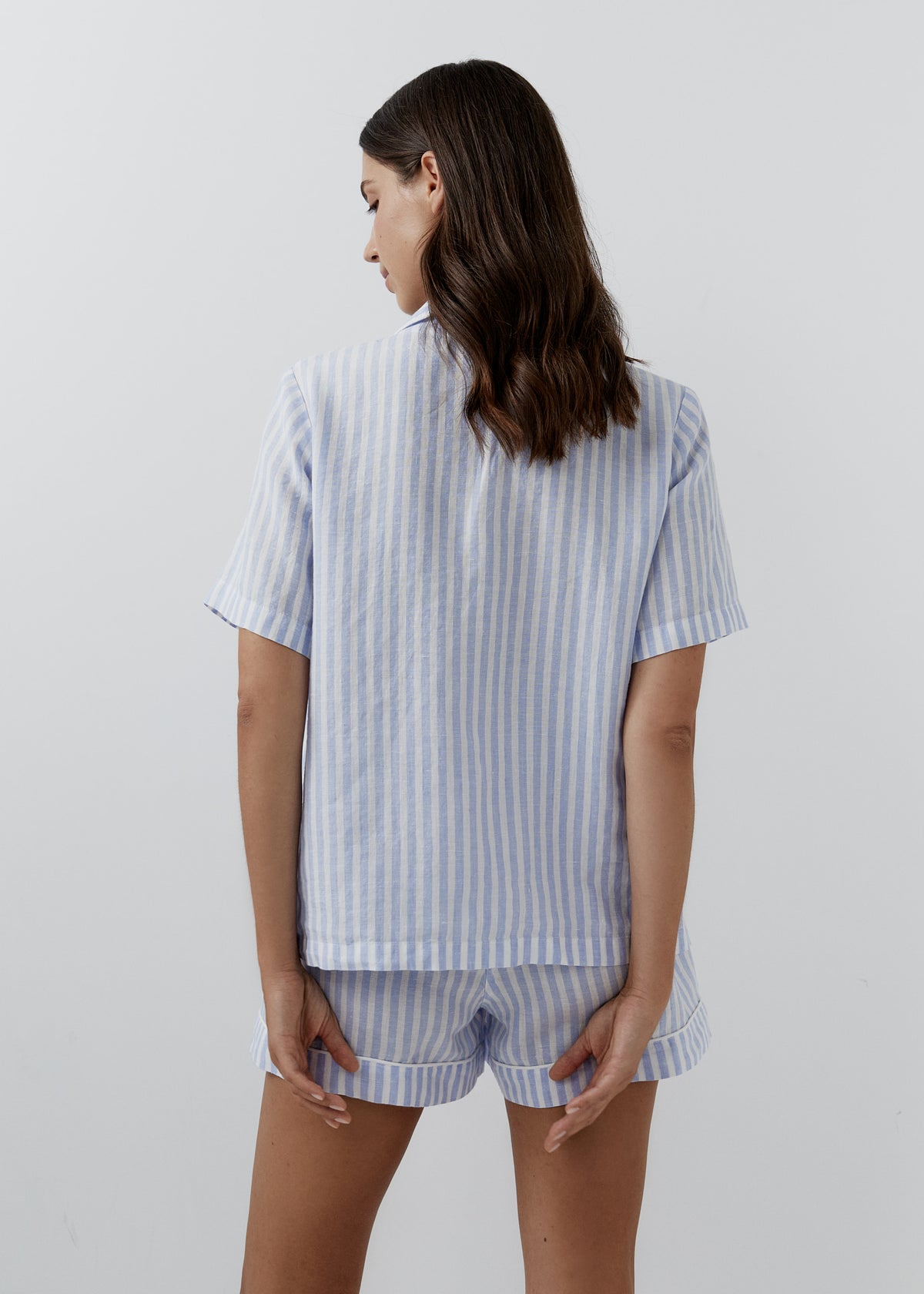 A woman shown from the back wearing a light blue and white vertically striped short-sleeve linen pajama top and matching linen shorts against a plain light gray studio background.