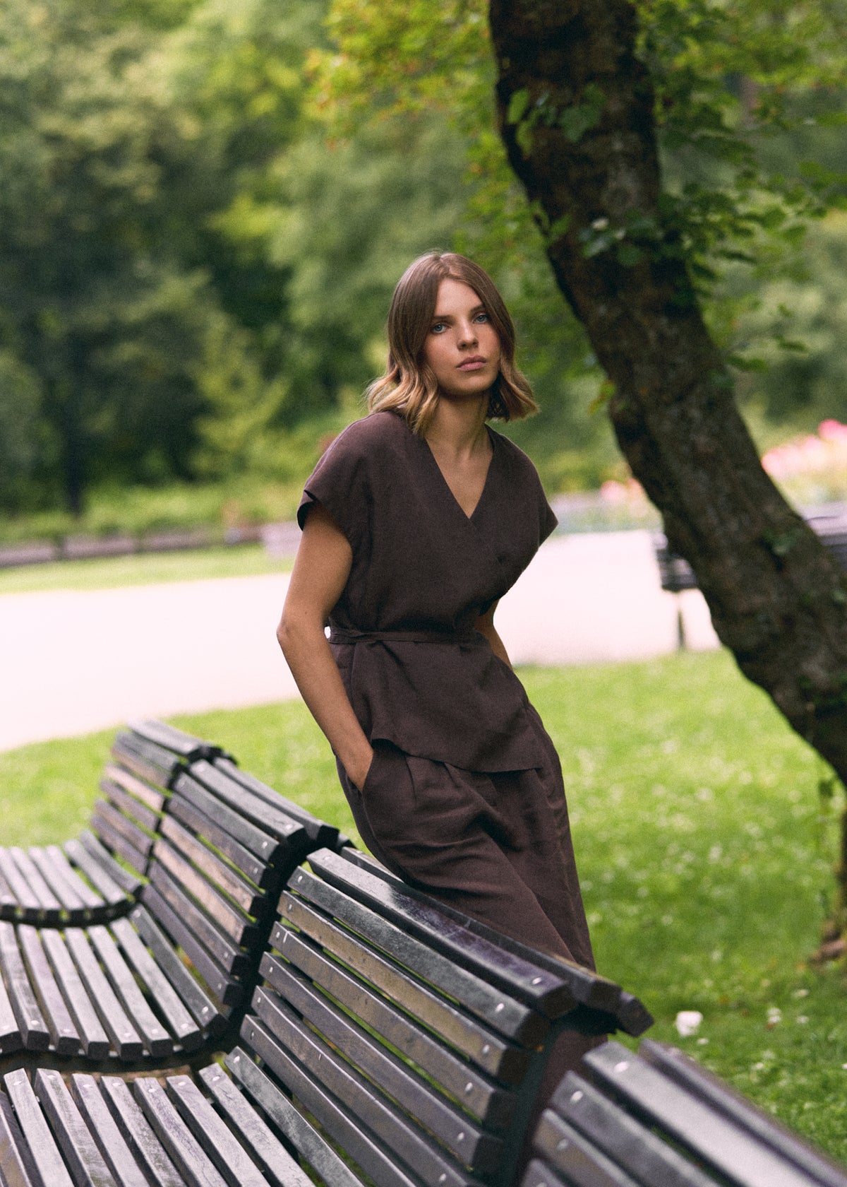 A woman wearing a dark brown short-sleeve wrap-style jumpsuit stands beside a park bench in a green outdoor setting.