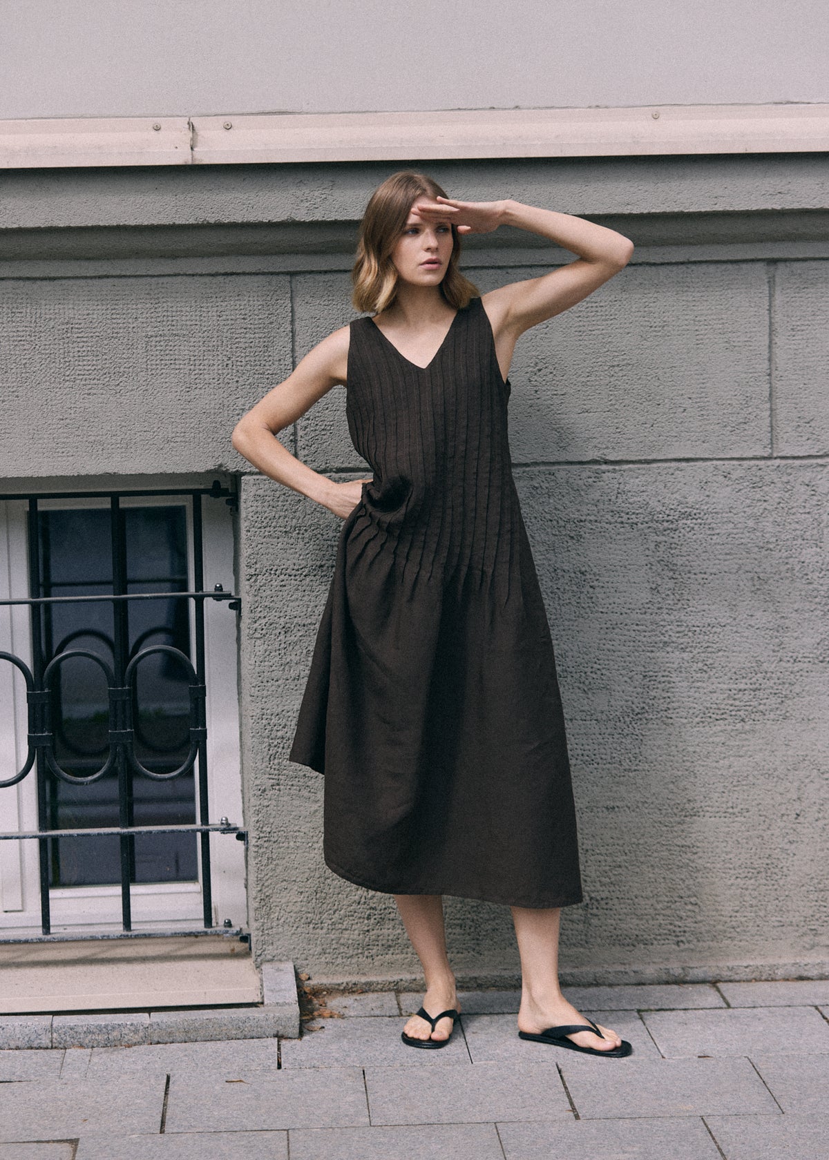 Woman wearing a dark brown sleeveless linen maxi dress with a V-neckline, vertical pleated detailing and a relaxed flowing skirt, standing against a grey urban wall.
