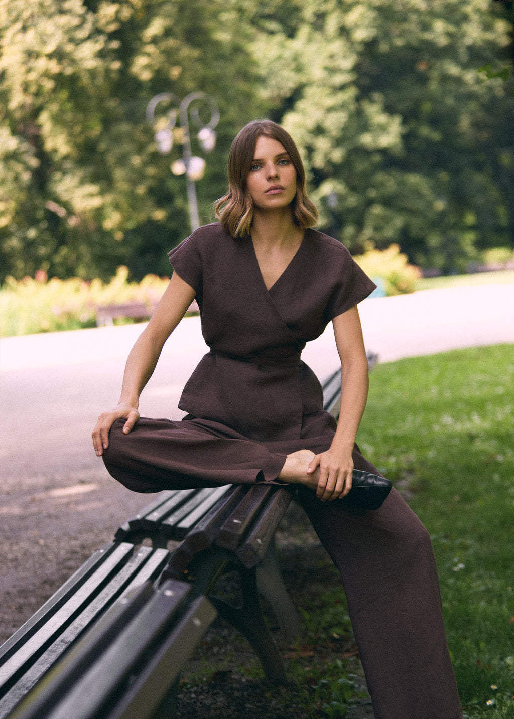 A woman wearing dark brown trousers and a matching wrap vest tied at the waist with a belt sits on a park bench with trees and green grass in the background.