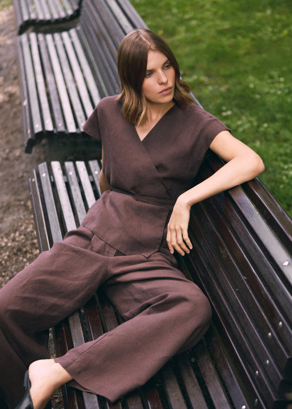 A woman wearing a matching brown wrap-style short-sleeve vest and wide-leg trousers sits on a dark wooden park bench with grass in the background.
