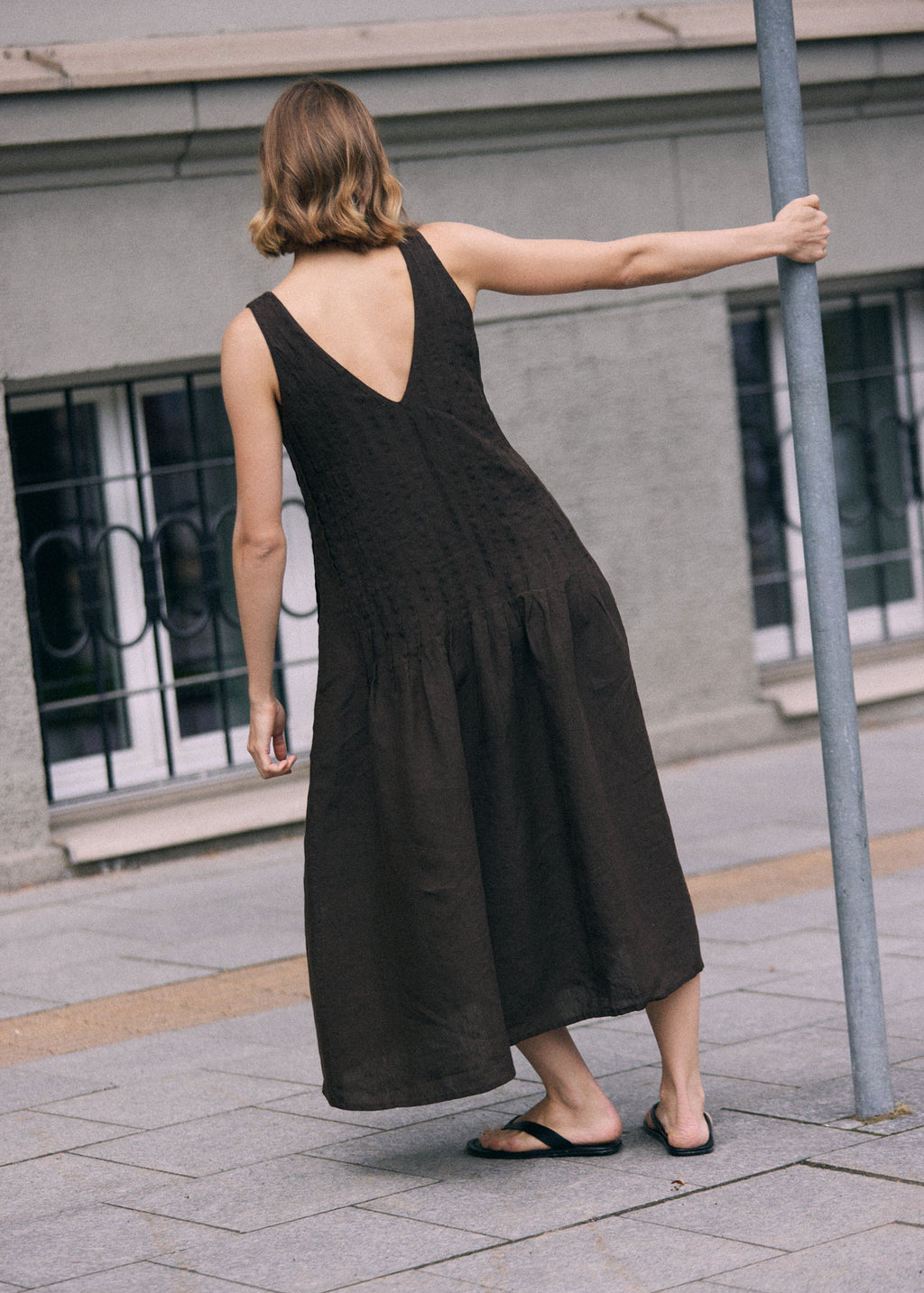 Woman photographed from behind wearing a dark brown sleeveless linen maxi dress with a deep V-shaped back, relaxed A-line silhouette and softly gathered lower skirt. The loose, ankle-length dress has a natural textured linen finish and subtle vertical detailing through the upper section. She wears simple black flat sandals and holds a metal pole with one hand while standing on a paved city sidewalk beside a grey building.
