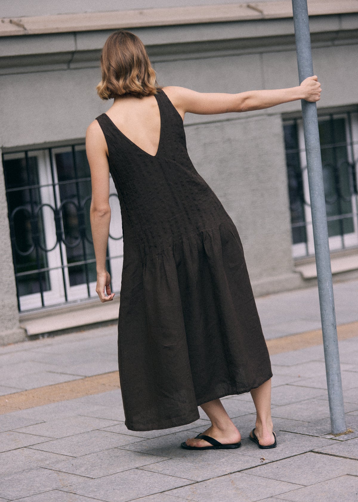 Woman photographed from behind wearing a dark brown sleeveless linen maxi dress with a deep V-shaped back, relaxed A-line silhouette and softly gathered lower skirt. The loose, ankle-length dress has a natural textured linen finish and subtle vertical detailing through the upper section. She wears simple black flat sandals and holds a metal pole with one hand while standing on a paved city sidewalk beside a grey building.
