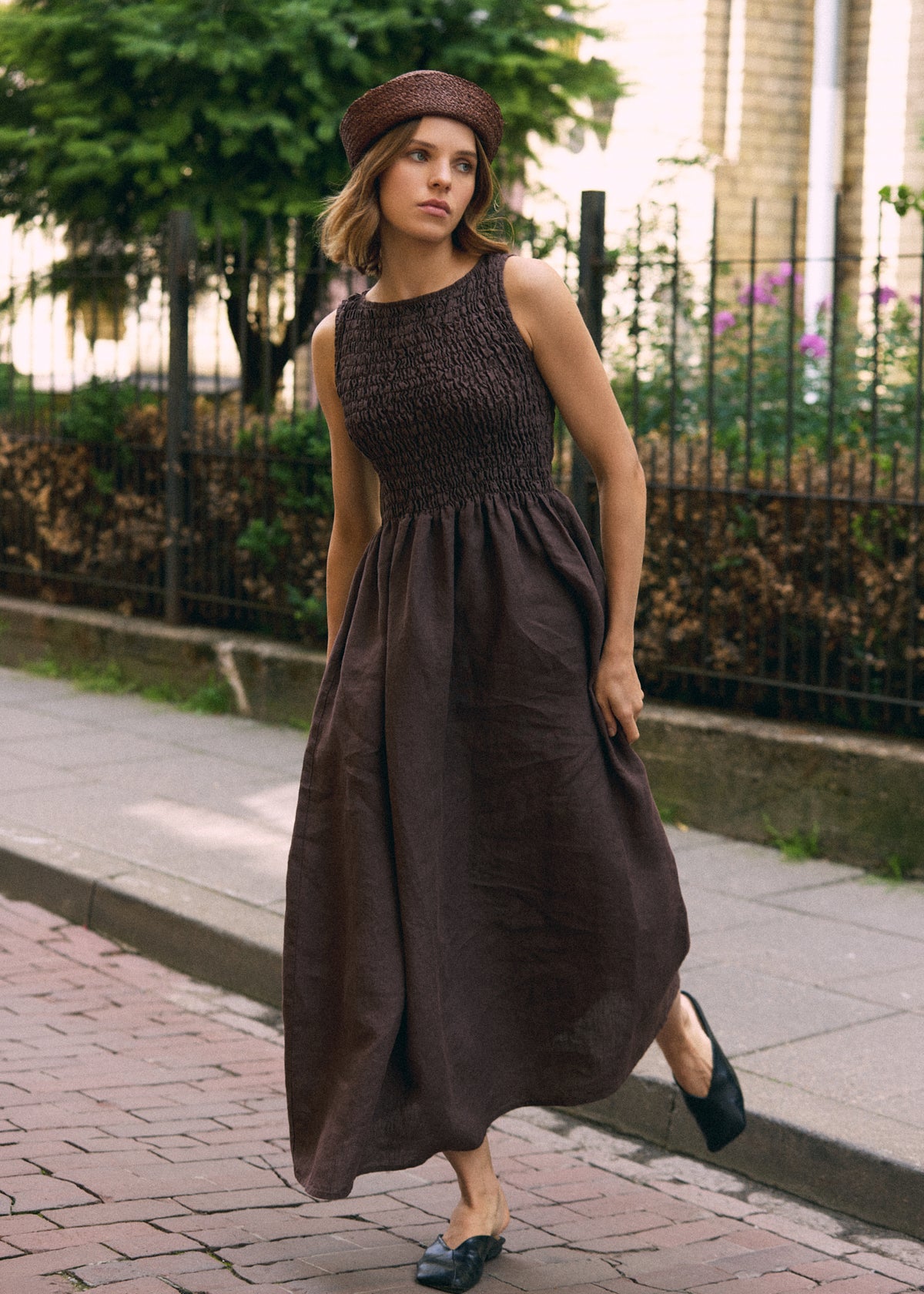 Woman walking in a dark brown sleeveless linen maxi dress featuring a textured shirred bodice, defined waist and long flowing skirt, paired with a brown woven hat and black flats.