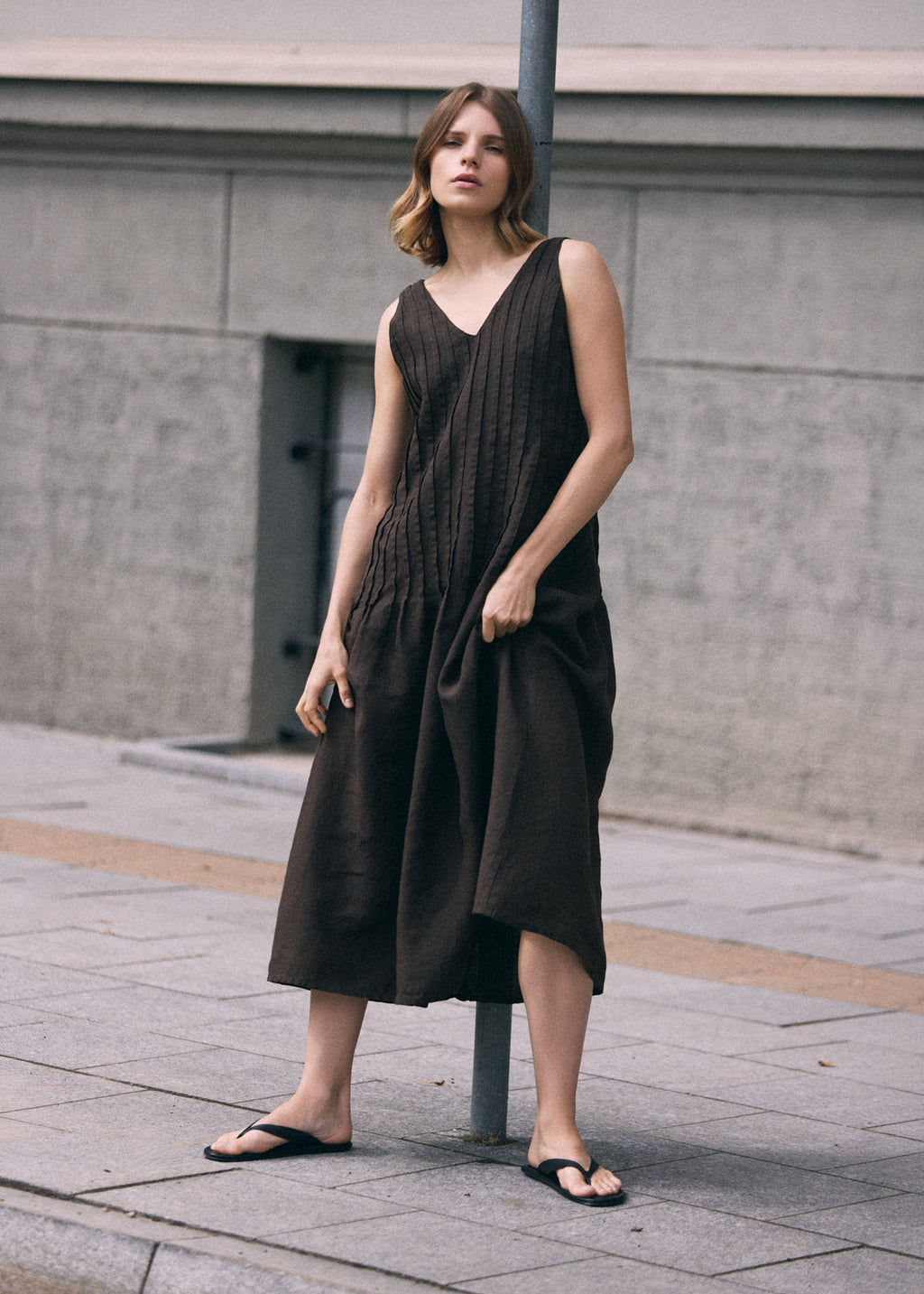 Woman posing beside a metal pole in a dark brown sleeveless linen maxi dress with a V-neckline, vertical pleated front and softly flowing skirt.