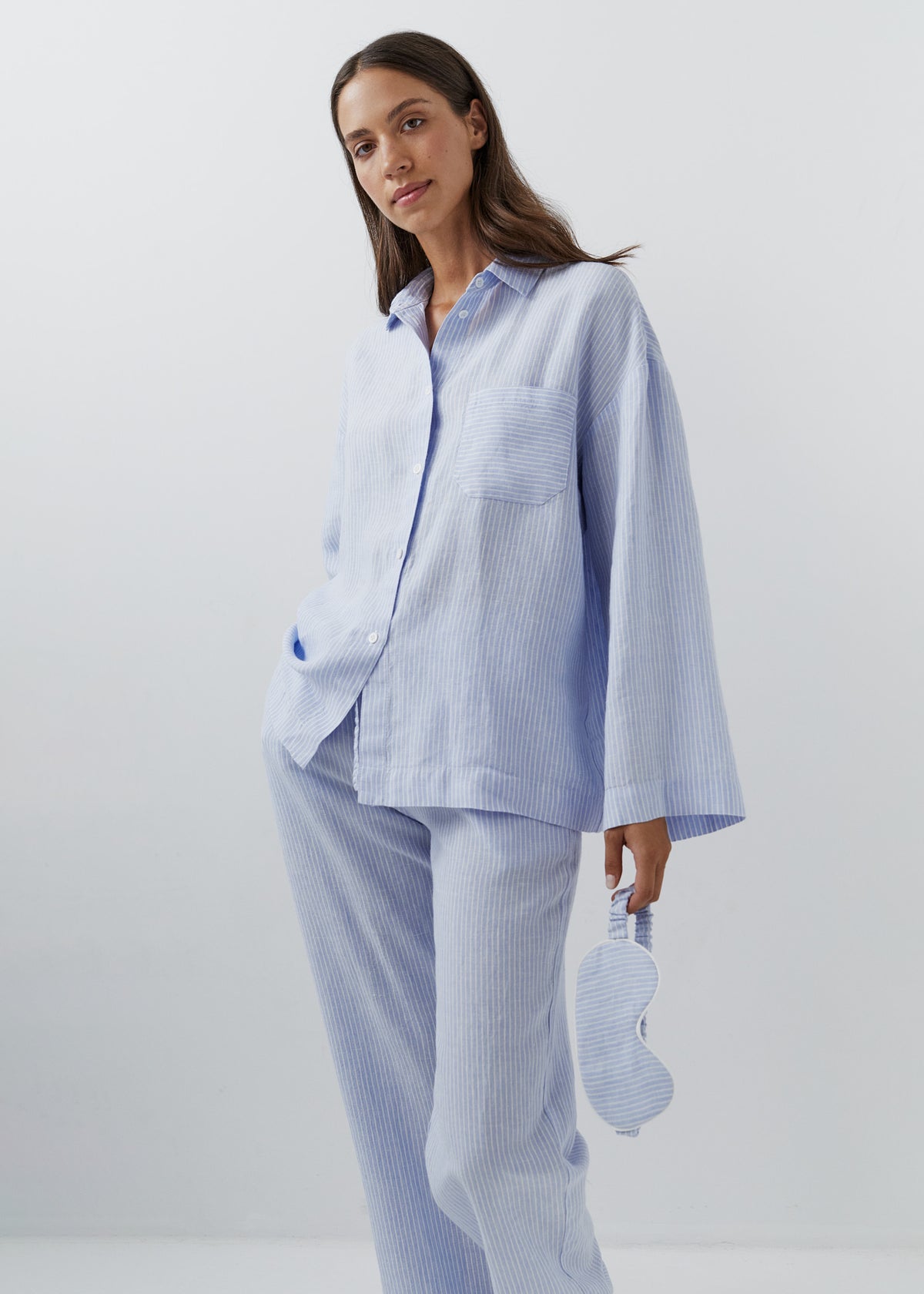 Woman wearing fine blue and white striped linen trousers with a matching linen shirt, holding a linen eye mask.
