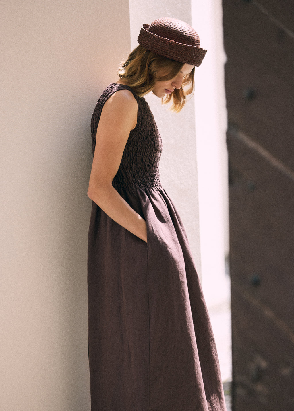 Side view of a woman wearing a dark brown linen maxi dress with a textured shirred bodice and side pocket, styled with a brown woven hat in soft natural sunlight.