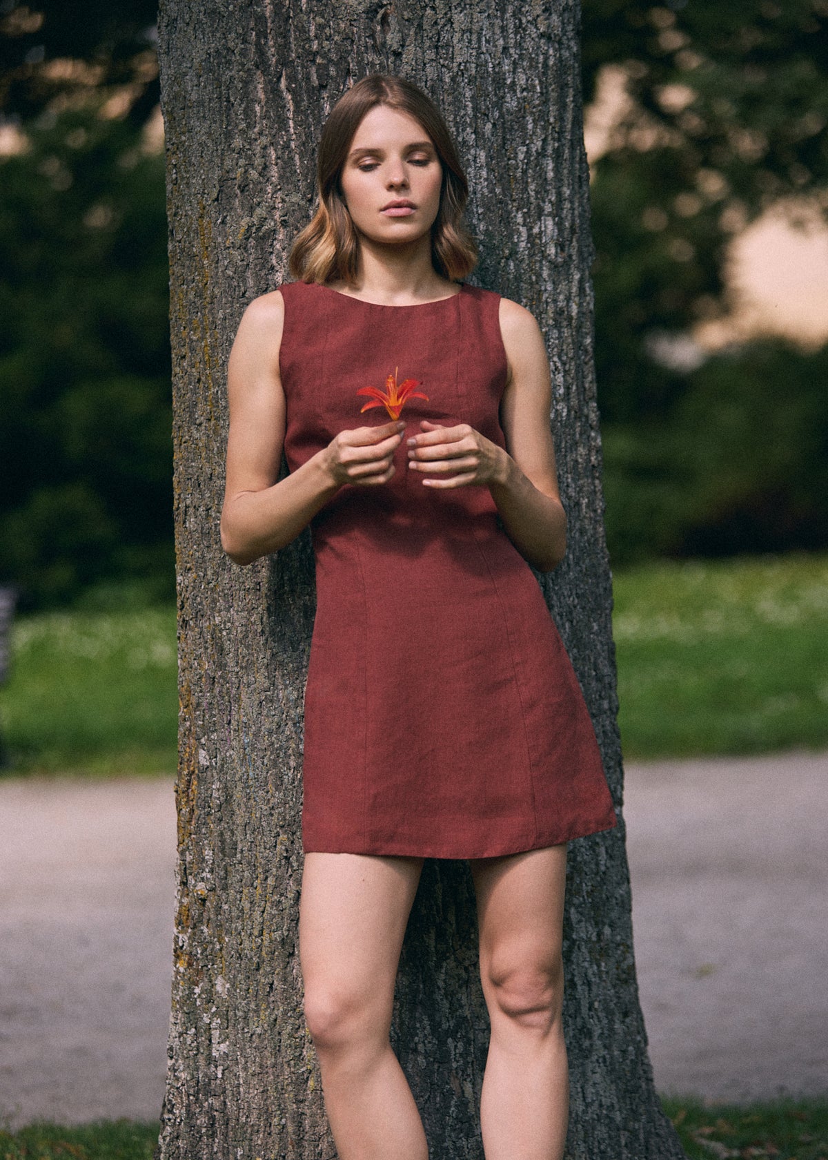 A woman wearing a sleeveless rust-red linen dress stands leaning against a large tree trunk in an outdoor park setting.