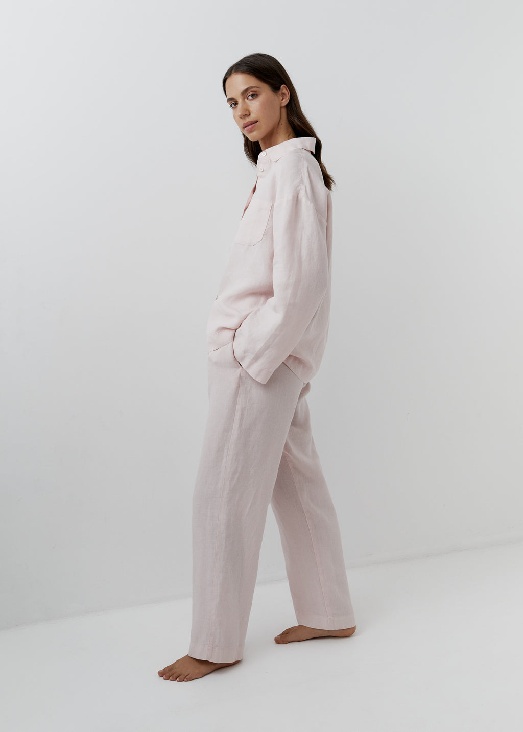 A woman modeling a light pink two-piece pajama set consisting of a long-sleeve button-up shirt and matching wide-leg pants, standing barefoot against a clean white studio background.