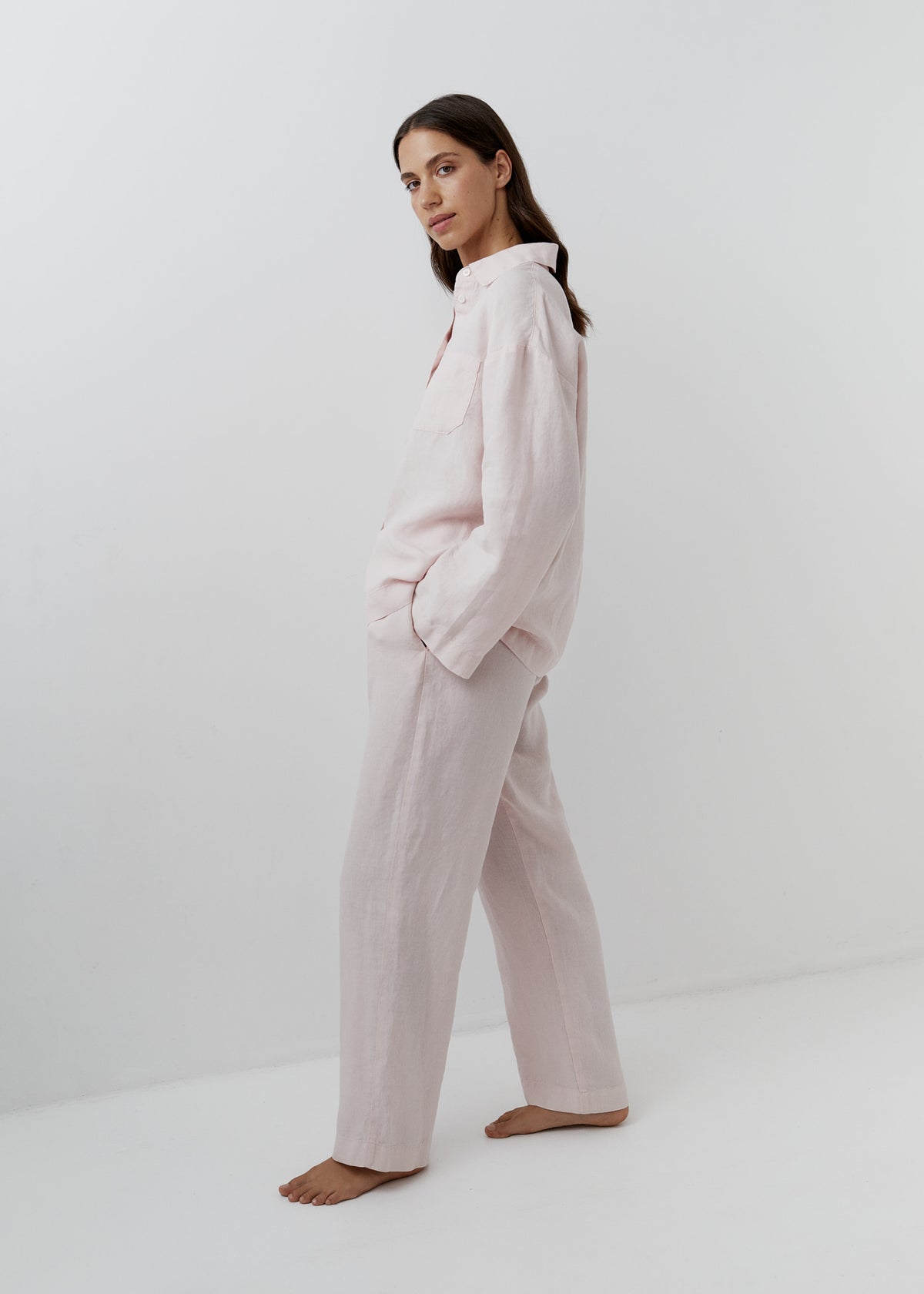 A woman modeling a light pink two-piece pajama set consisting of a long-sleeve button-up shirt and matching wide-leg pants, standing barefoot against a clean white studio background.