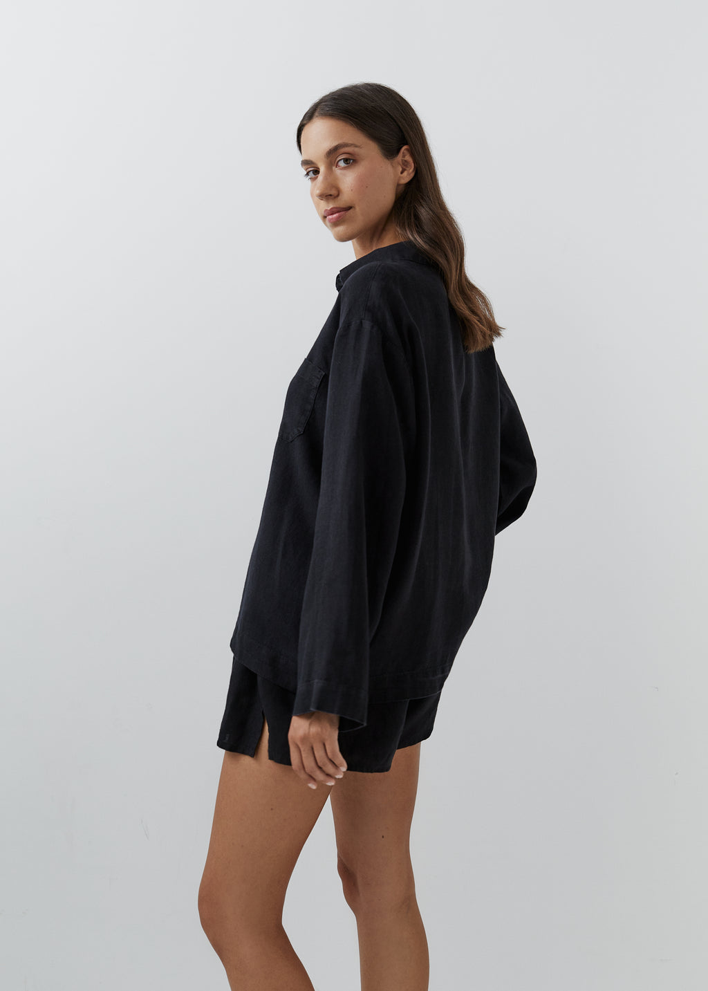 A woman modeling an oversized black long-sleeve shirt with matching black shorts against a plain light gray studio background.