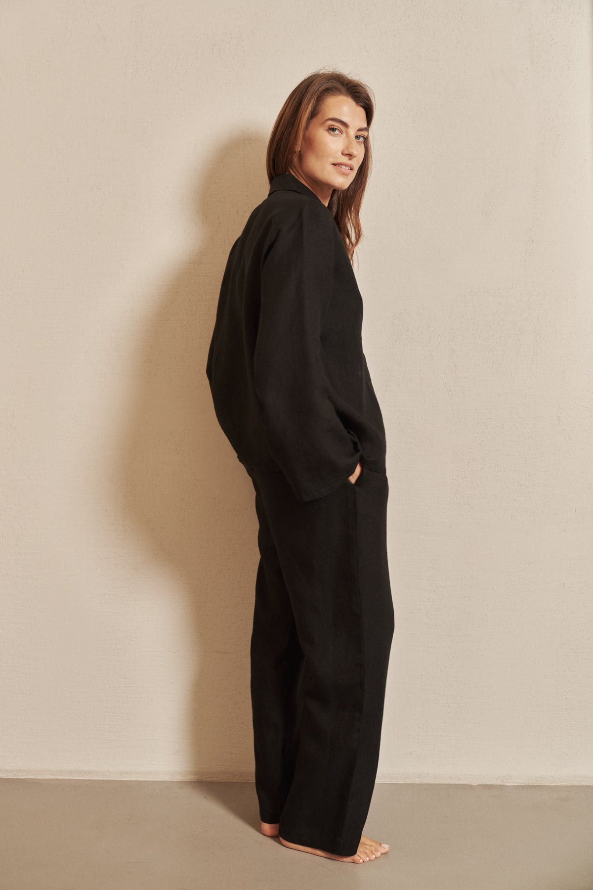 A woman stands barefoot against a plain beige wall, wearing a loose black long-sleeve top and matching wide-leg pants.
