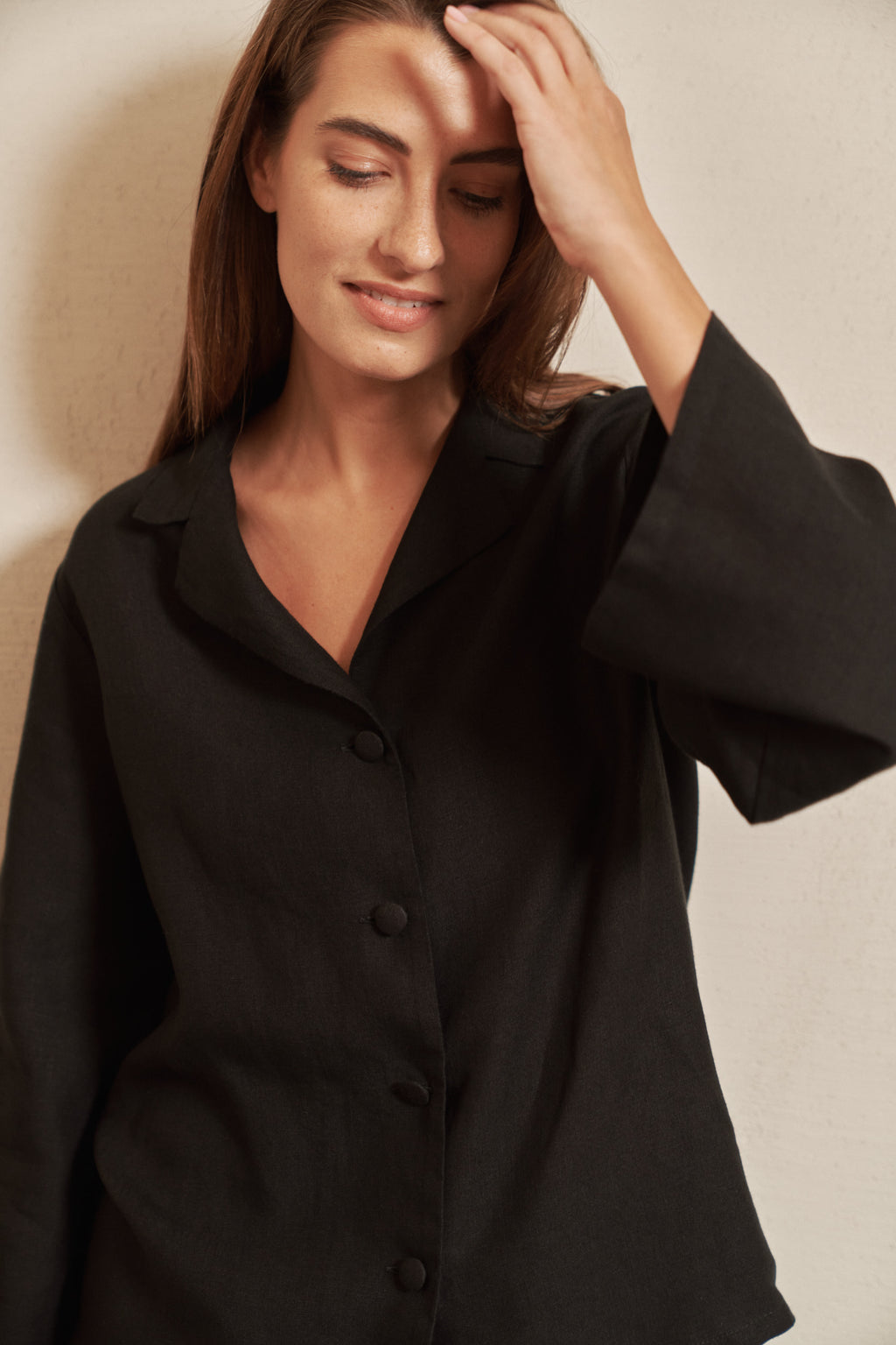 A woman wearing a black button-front shirt jacket with wide sleeves, posed against a neutral beige wall.