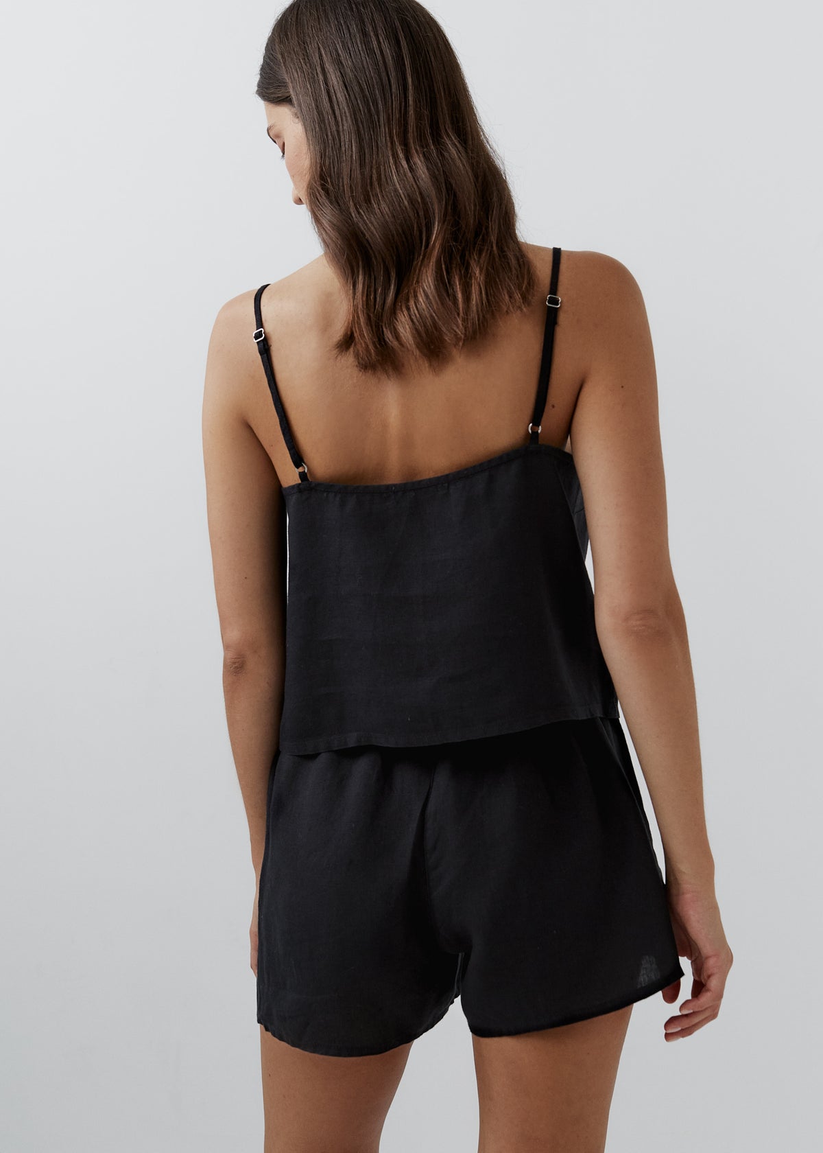 A woman shown from the back wearing a black camisole-style romper with thin shoulder straps and matching shorts against a plain light gray studio background.