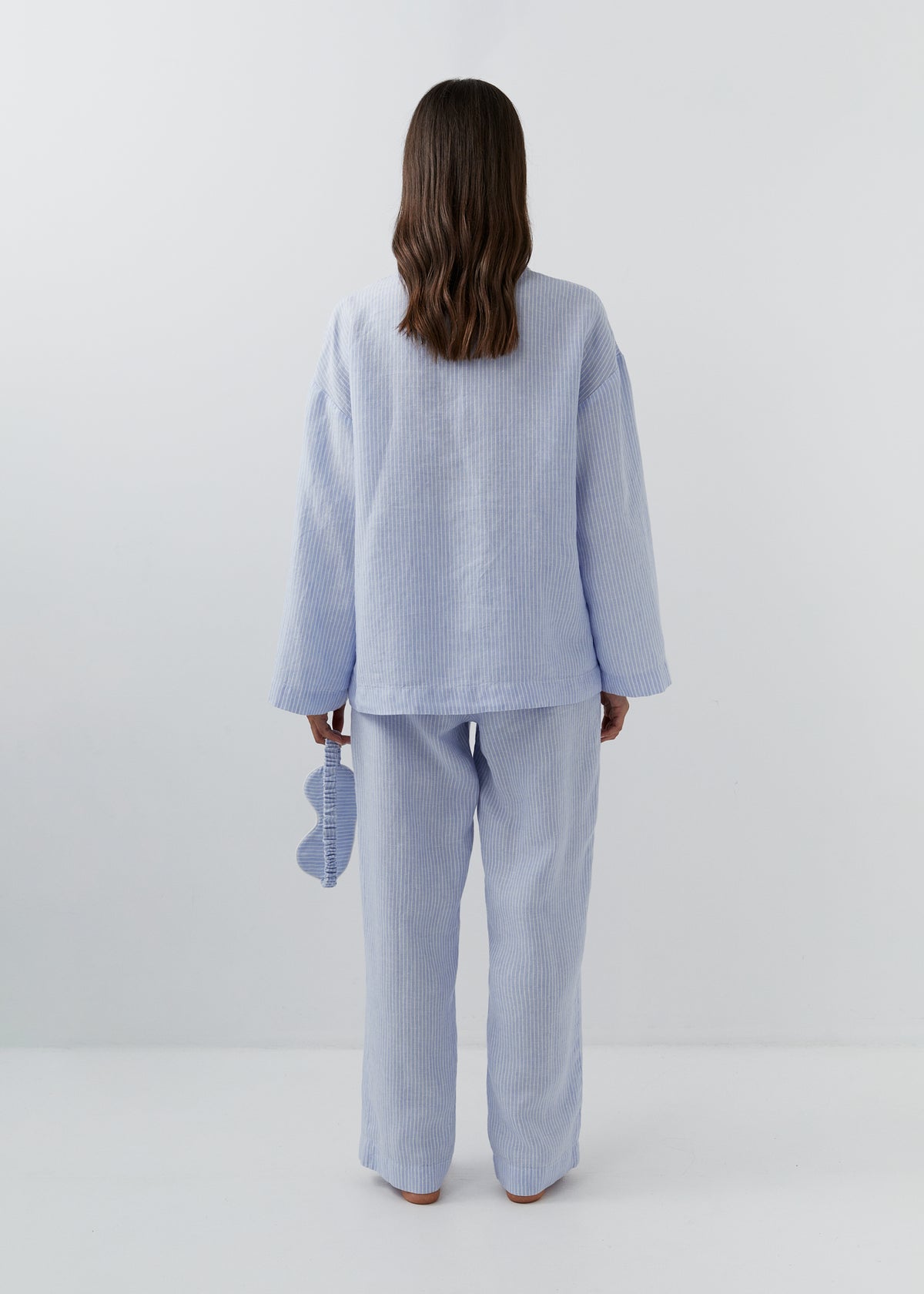 A woman stands with her back to the camera wearing a light blue linen sleepwear set consisting of a long-sleeve top and matching wide-leg pants. She is holding a matching light blue pillow in her right hand, set against a clean white studio background.
