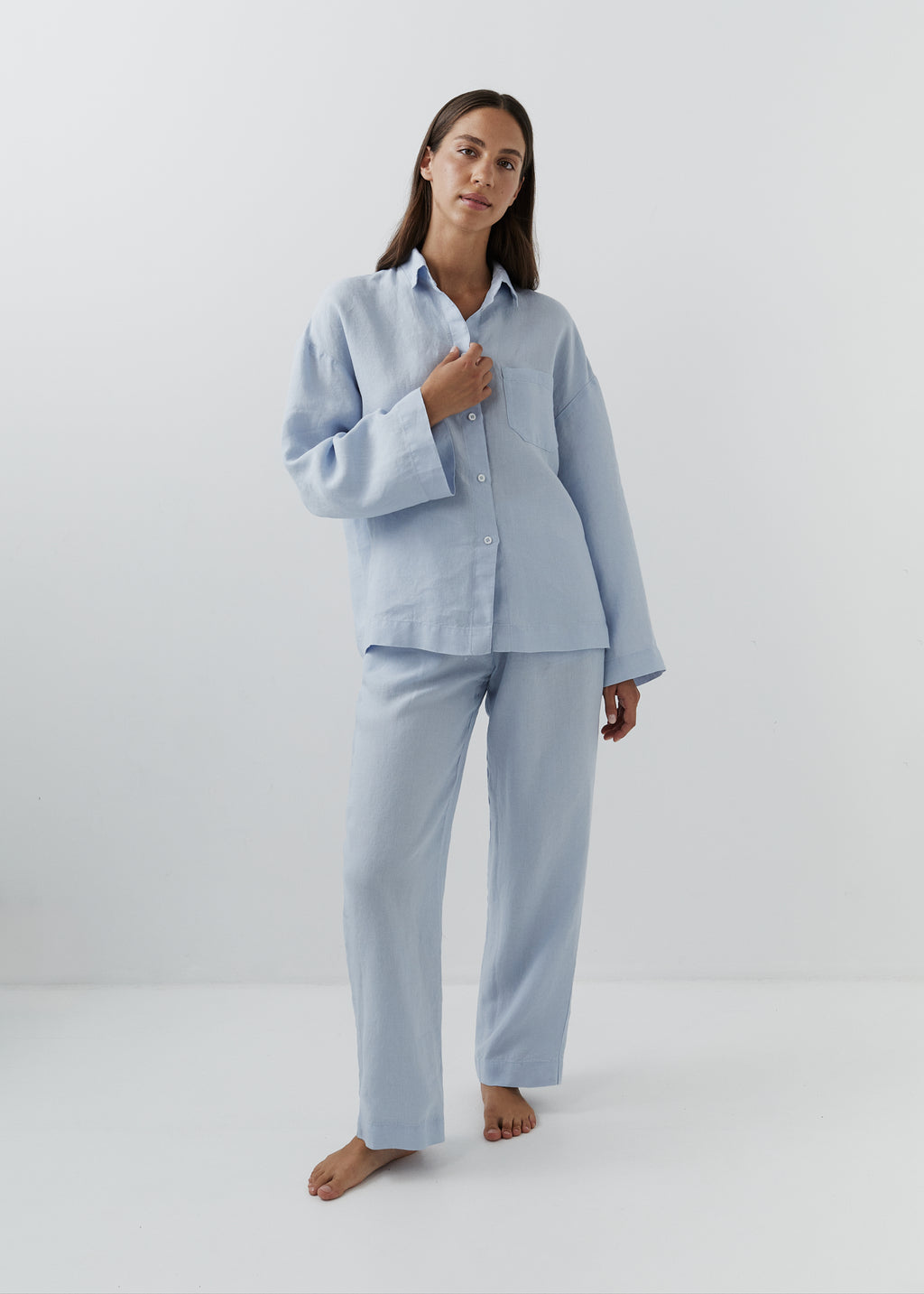 A woman modeling a light blue loungwear set consisting of a long-sleeve button-up shirt and matching wide-leg pants, standing against a plain white studio background.