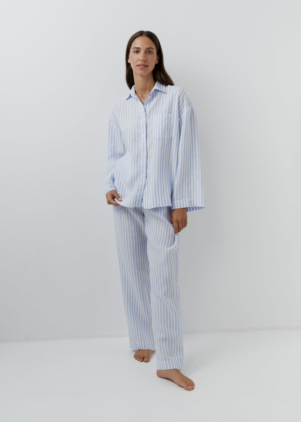 A woman modeling a light blue and white vertically striped pyjama set, consisting of a long-sleeve button-up shirt and matching trousers, standing against a plain white studio background.