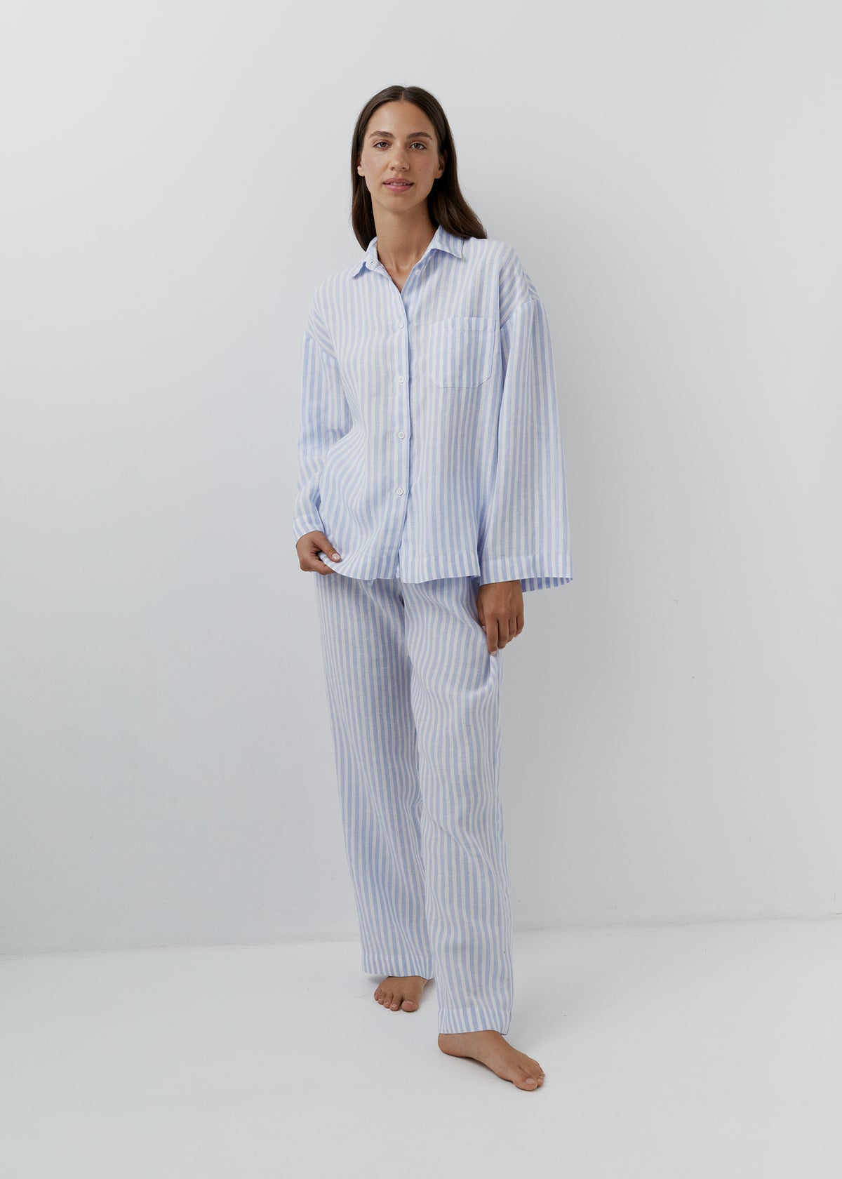 A woman modeling a light blue and white vertically striped pyjama set, consisting of a long-sleeve button-up shirt and matching trousers, standing against a plain white studio background.