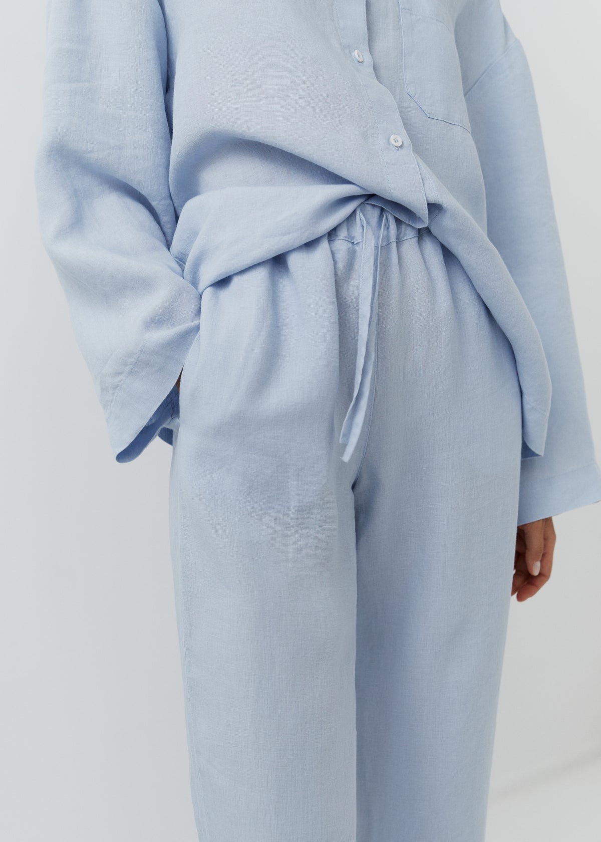 A close-up of a person wearing a light blue linen two-piece set consisting of a long-sleeve button-front shirt and matching wide-leg trousers with a drawstring waist.