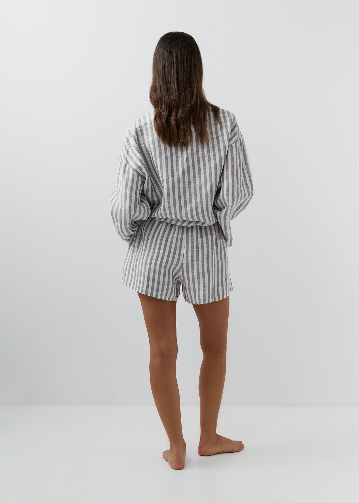 A woman viewed from behind wearing a matching gray and white vertically striped lounge set consisting of a long-sleeve top and shorts, standing barefoot against a plain white studio background.