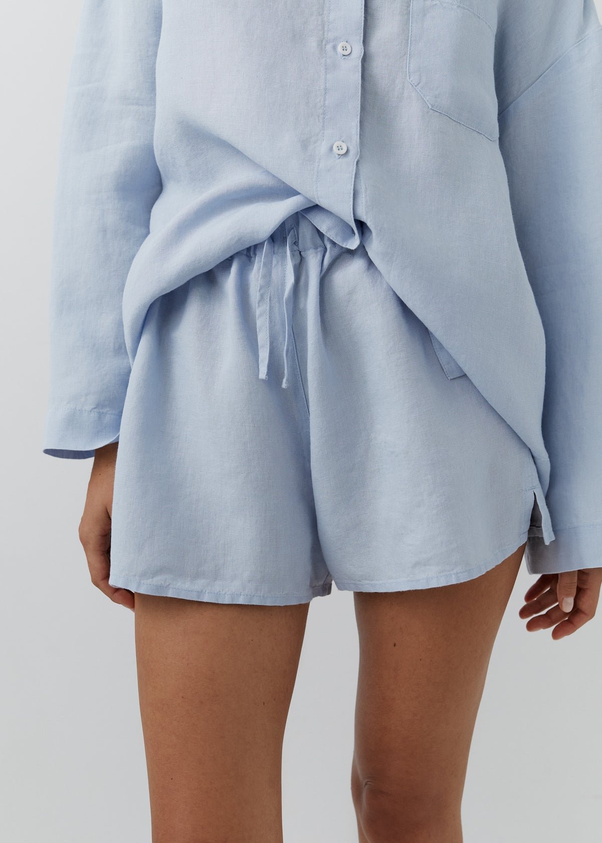 A close-up of a model wearing a light blue linen two-piece set consisting of a long-sleeve button-up shirt and matching shorts with a drawstring waist.