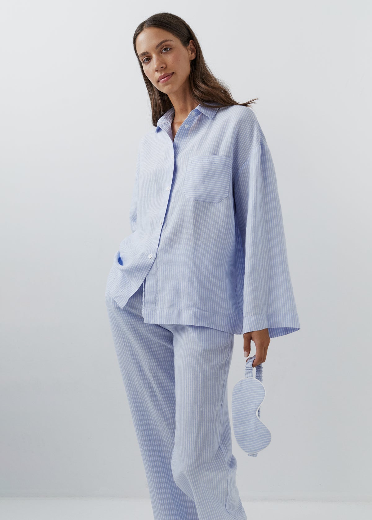 A woman modeling a light blue and white striped 100% linen pajama set, featuring a long-sleeve button-up shirt and matching pants, while holding a matching striped sleep mask against a plain light gray background.
